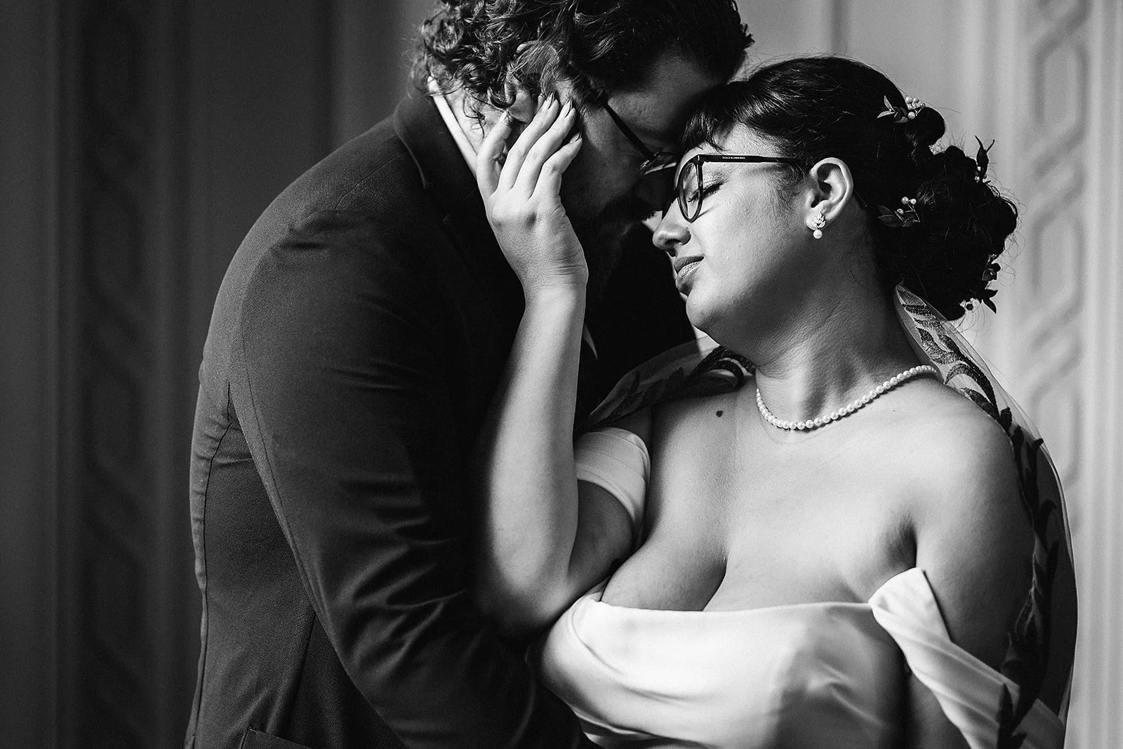 A black and white photograph of a couple in an intimate moment, with their foreheads and noses touching, eyes closed. The woman is wearing glasses, pearl earrings, a pearl necklace, and an off-the-shoulder dress. The man has curly hair, glasses, and 
