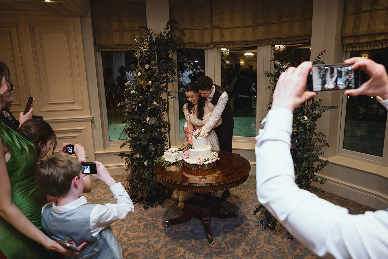 A newlywed couple cutting their wedding cake surrounded by family and friends, some taking photos.