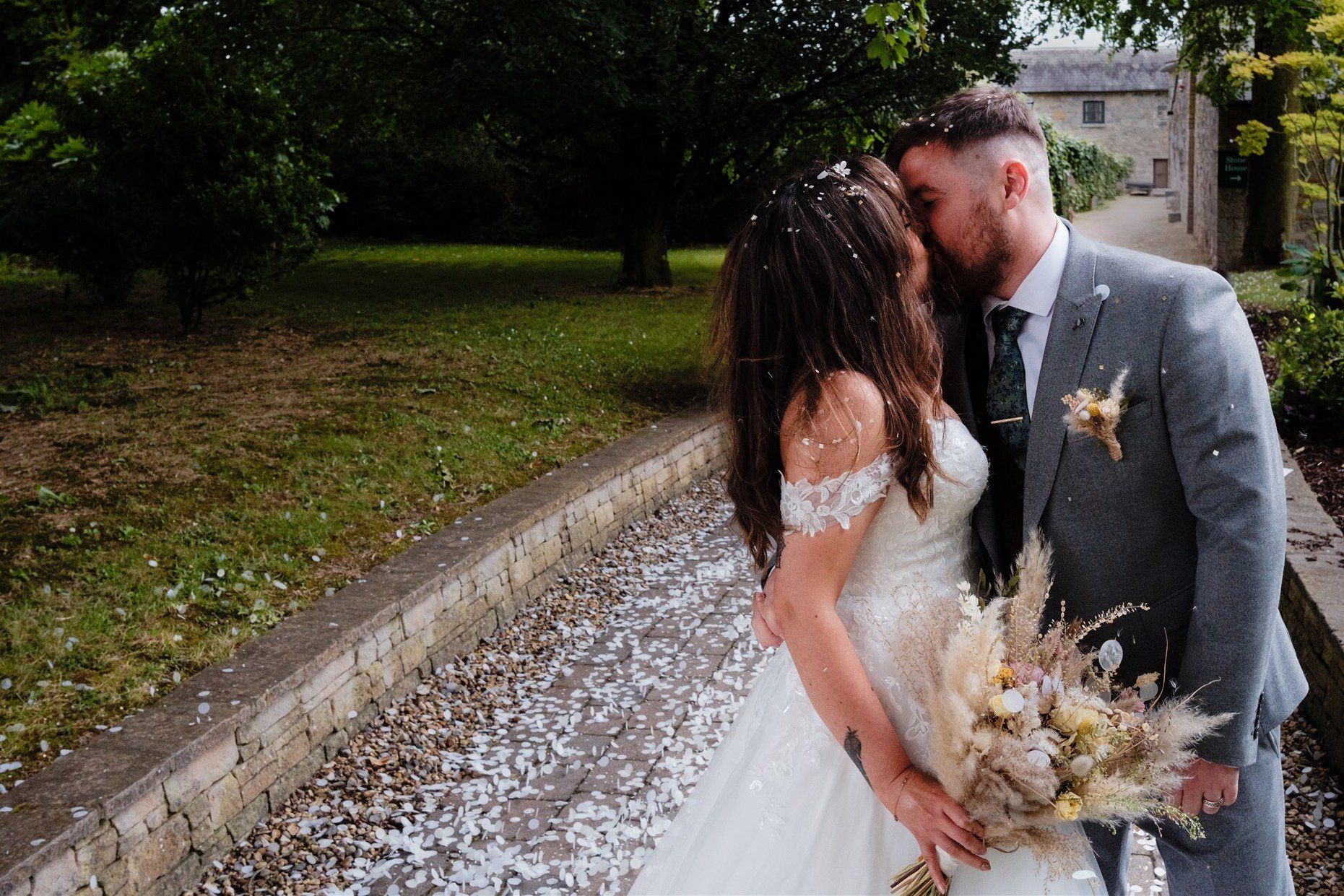 A bride and groom share a kiss at their wedding outdoors, with flower petals scattered on the path and trees in the background.