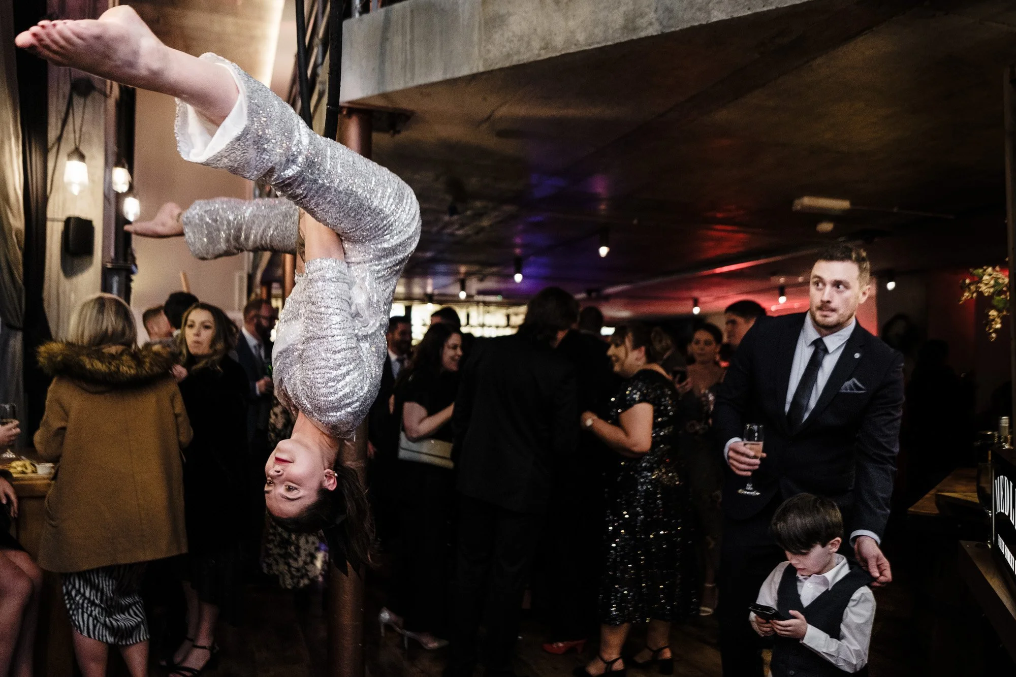 A girl in a shiny silver dress is performing a handstand at a crowded indoor event or party, while people around her are engaged in conversations and looking at their phones.