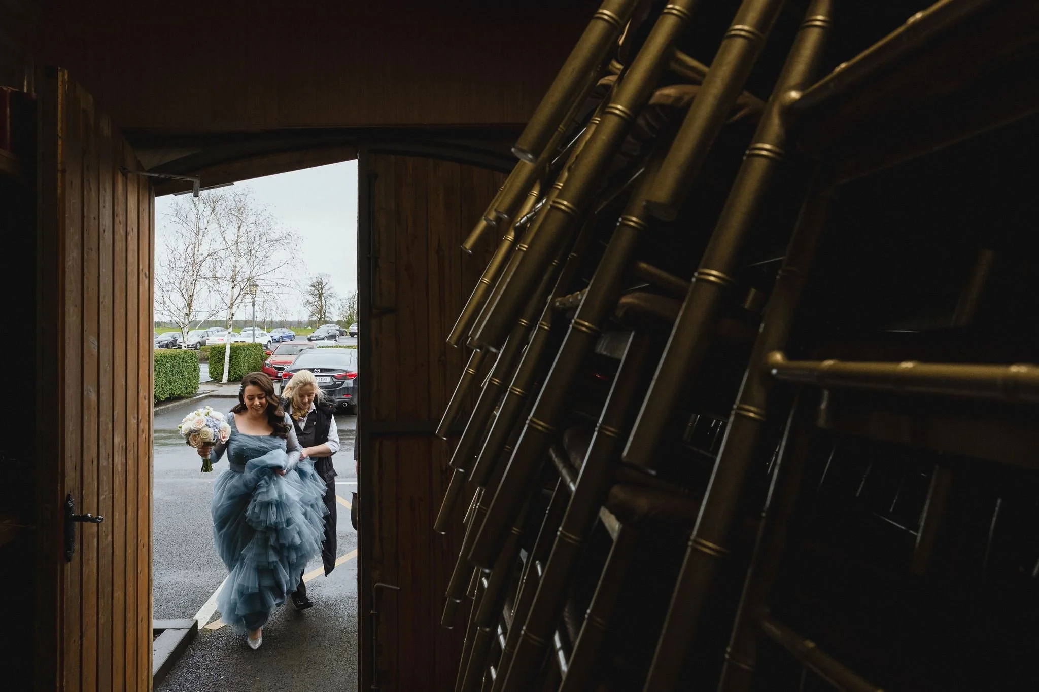 Bride holding a bouquet walking with a woman in a black jacket outside a building with chairs stacked inside visible through an open door.