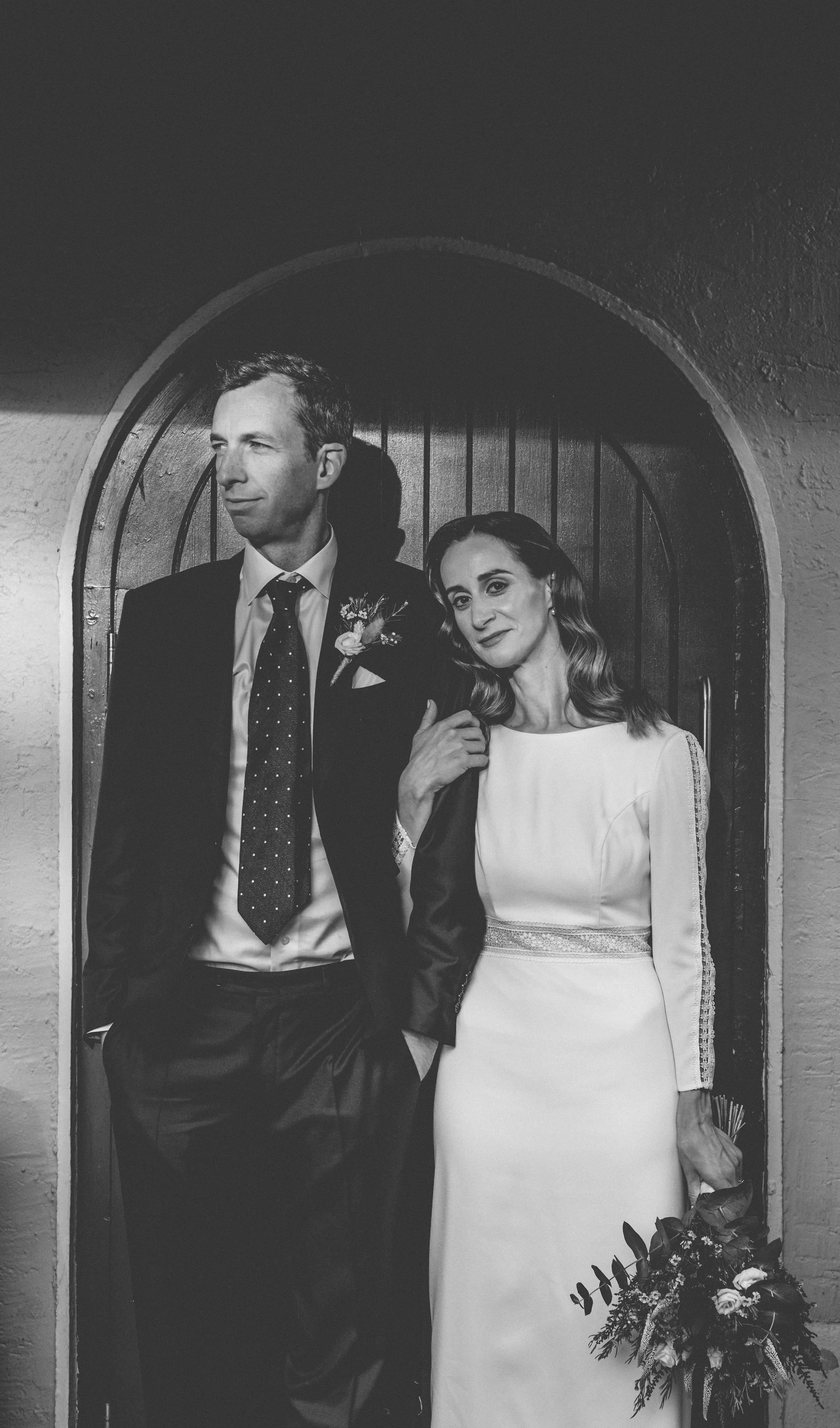 A black and white photo of a man and woman dressed in formal attire, standing in front of an arched wooden door. The man is wearing a suit with a boutonniere and the woman is in a wedding dress holding a bouquet of flowers.