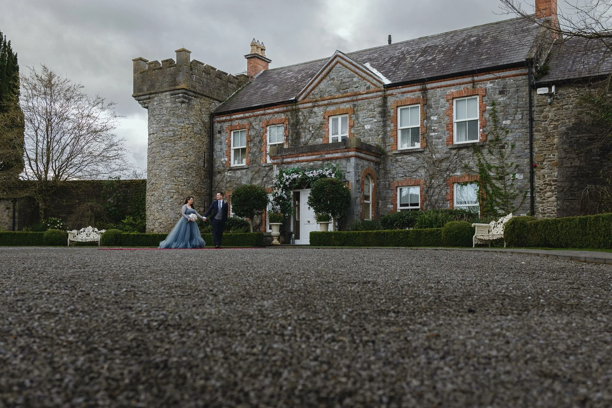 A bride and groom holding hands outside a stone mansion with a tower, decorated with white flowers and greenery, on an overcast day.