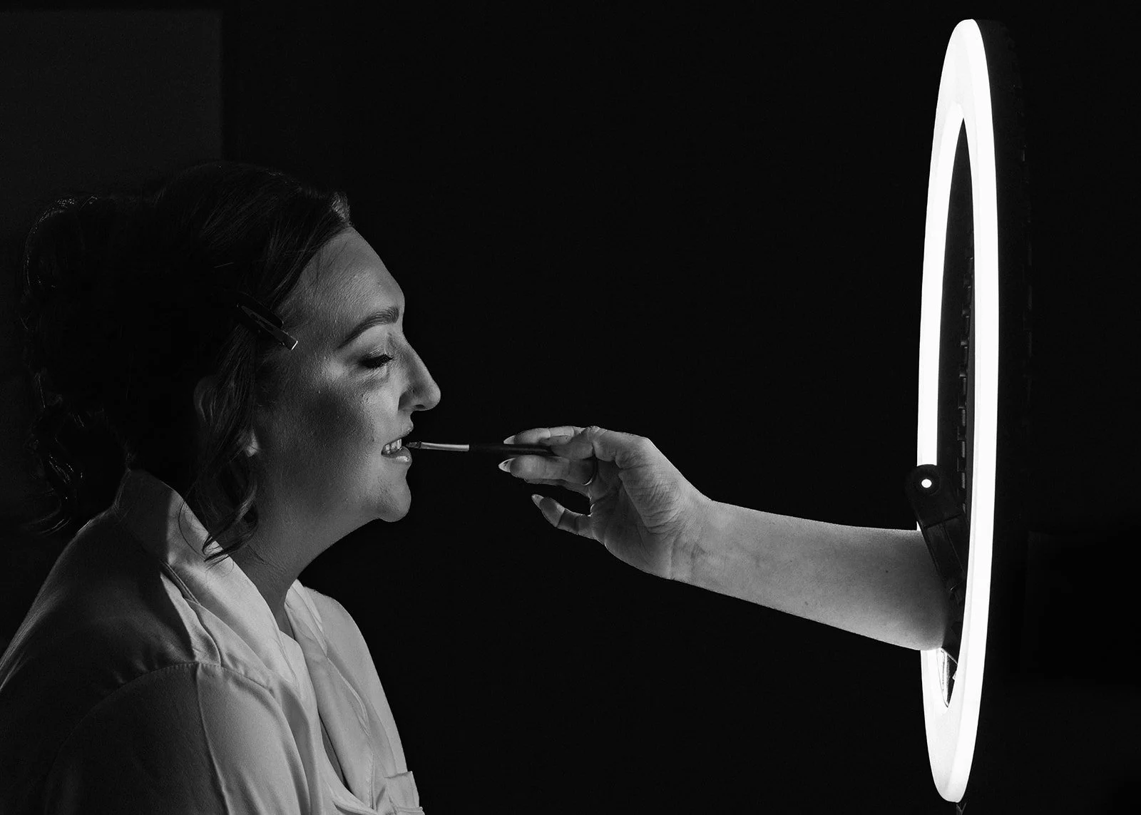 A woman with dark hair styled in waves is receiving a cosmetic or medical treatment from an unseen practitioner. The practitioner's hand is holding a small tool near the woman's mouth, projected through a circular lighted device, creating a professio