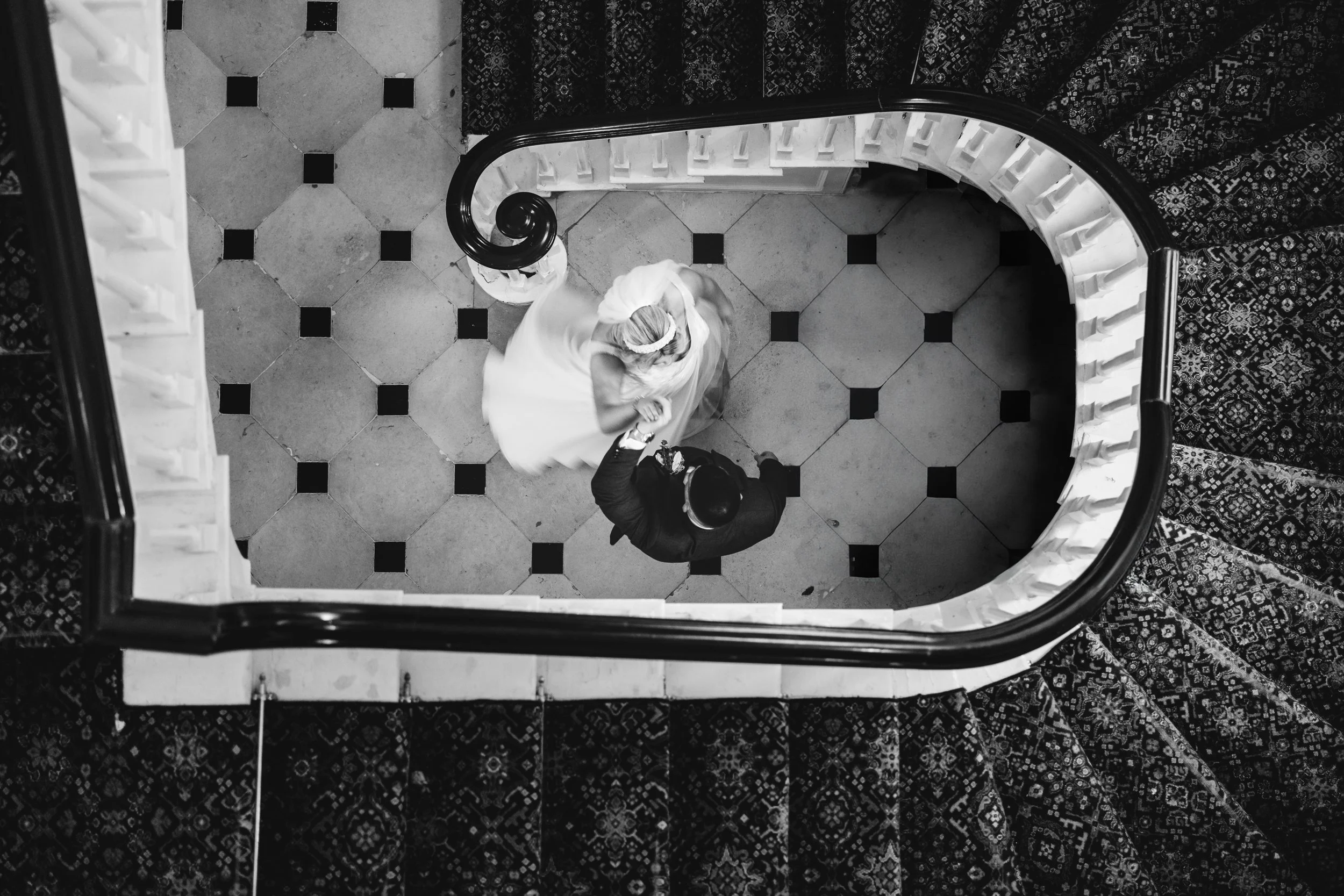 A black and white photo of a couple dancing on a tiled floor at the bottom of a spiral staircase, viewed from above.