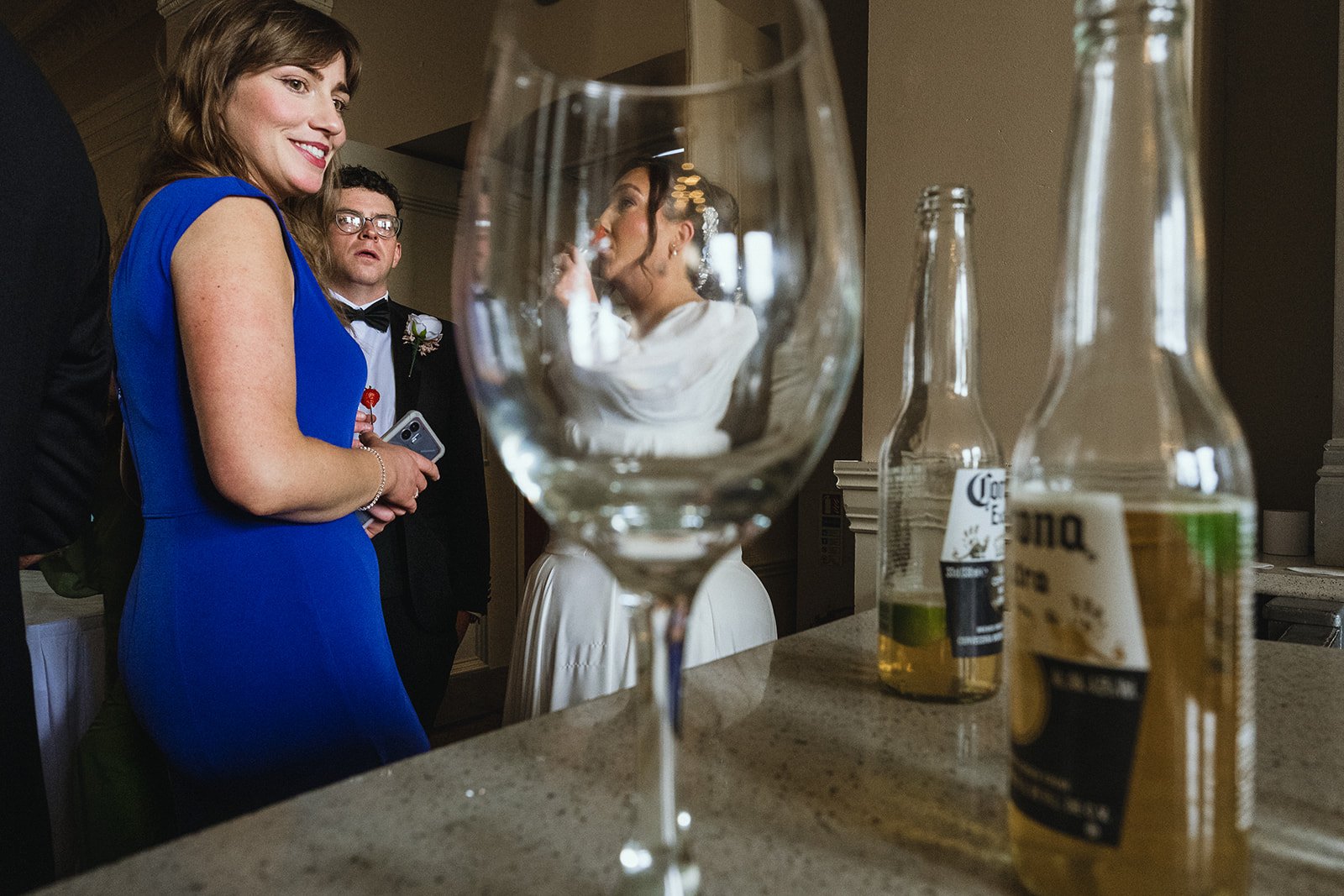 A group of people at a social gathering or party, with a focus on a woman in a bright blue dress smiling, a man in a tuxedo, and a woman in a white dress. In the foreground, an empty wine glass and some bottles of beer are visible on a countertop.