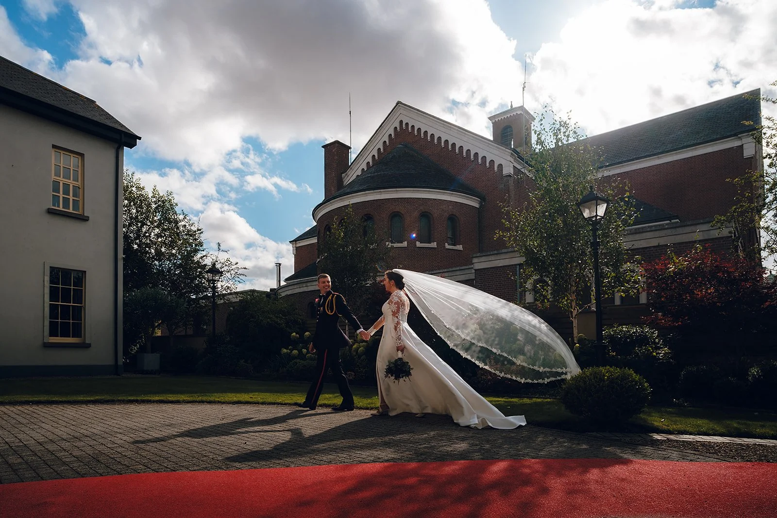 A bride and groom walking hand in hand outside a church, with the bride wearing a long white dress and veil, and the groom in a dark military uniform, under a partly cloudy sky.