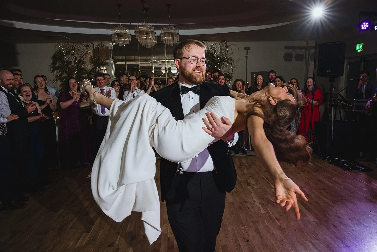 A groom in a tuxedo with a bowtie dances with a bride in a white wedding dress at their wedding reception, with guests in the background clapping and smiling.