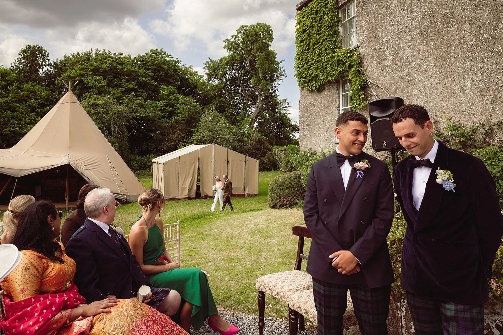 Two men in tuxedos with boutonnieres stand smiling and looking at each other at a wedding celebration outdoors, with guests seated and a bride and groom walking toward a tent in the background.