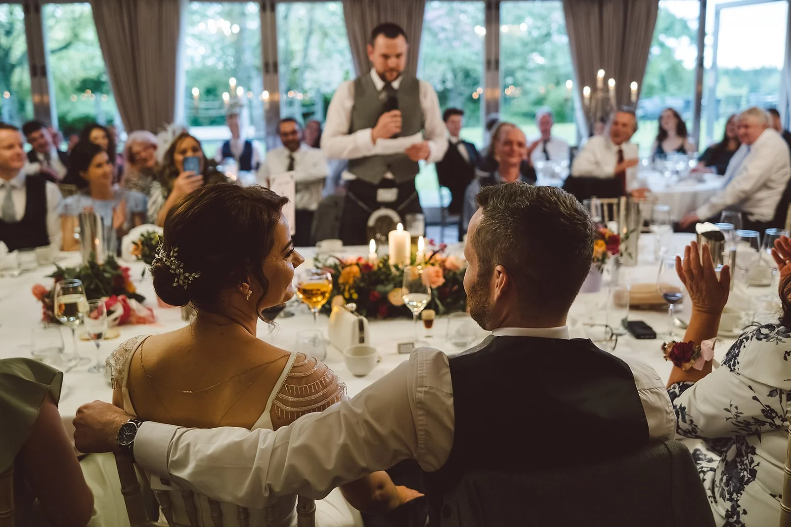A wedding reception with a bride and groom sitting at a table, surrounded by guests, while a man gives a speech holding a microphone. The room has natural light and decorated with candles and flowers.