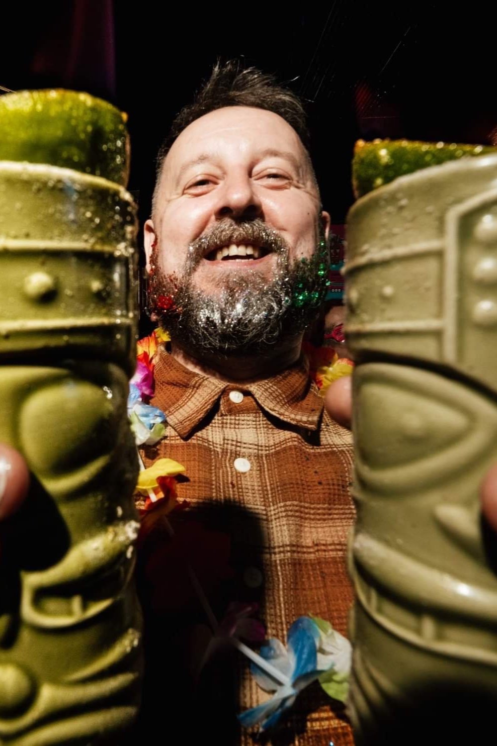 Man with beard and glitter, holding two green decorative mugs, wearing a plaid shirt and a colorful lei.