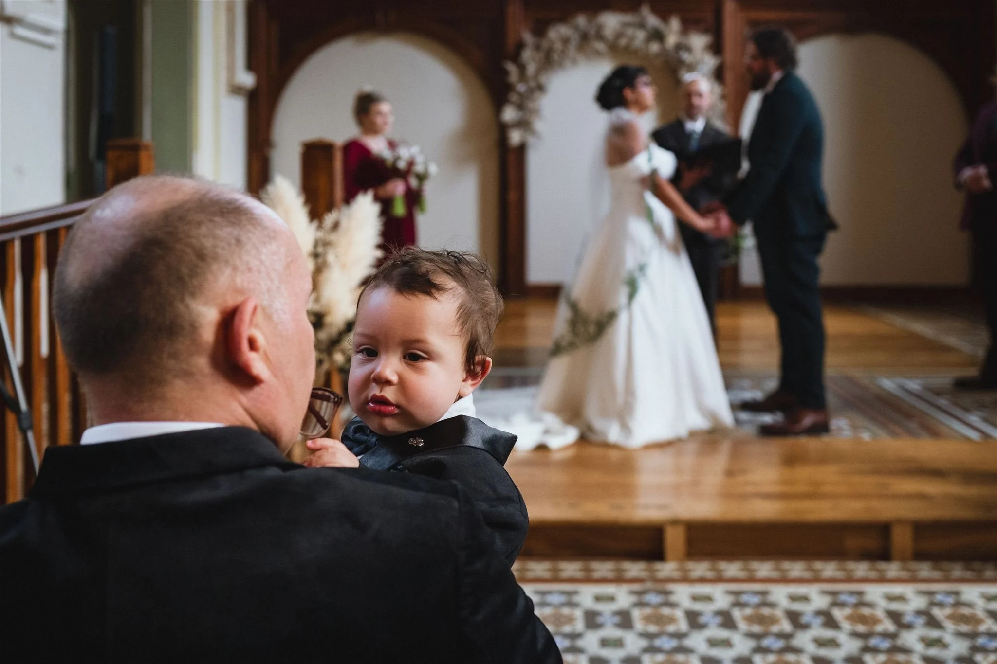 A man holding a young child during a wedding ceremony, with the bridal couple exchanging vows in the background.