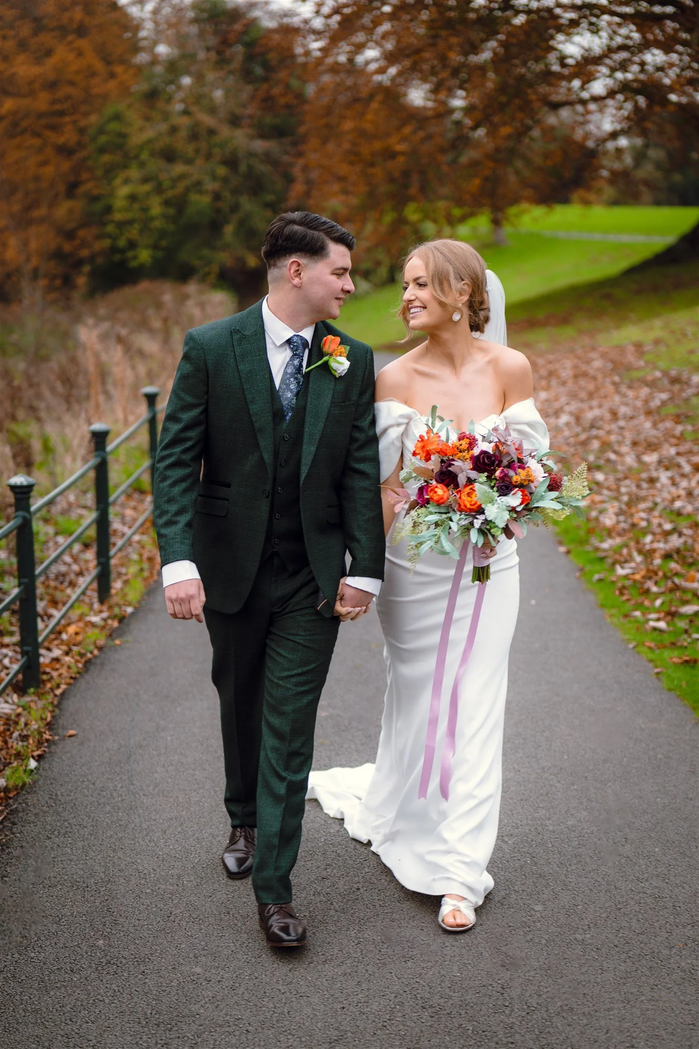 A newlywed couple walking outdoors in a park during autumn. The groom is in a dark green suit with a boutonniere, and the bride is in a white off-the-shoulder wedding dress holding a large bouquet of flowers. They are smiling and looking at each othe