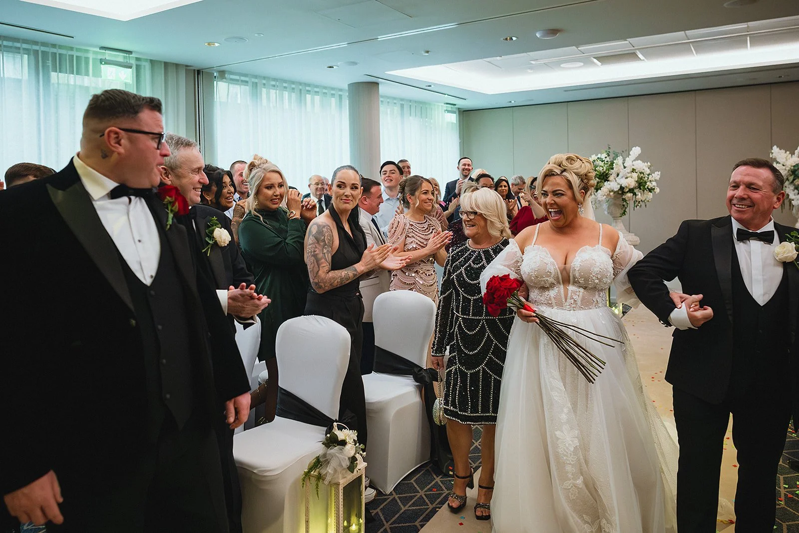 Bride in a white wedding gown holding a bouquet of red roses, walking down the aisle with groom in a black tuxedo, surrounded by guests clapping and smiling at a wedding ceremony.