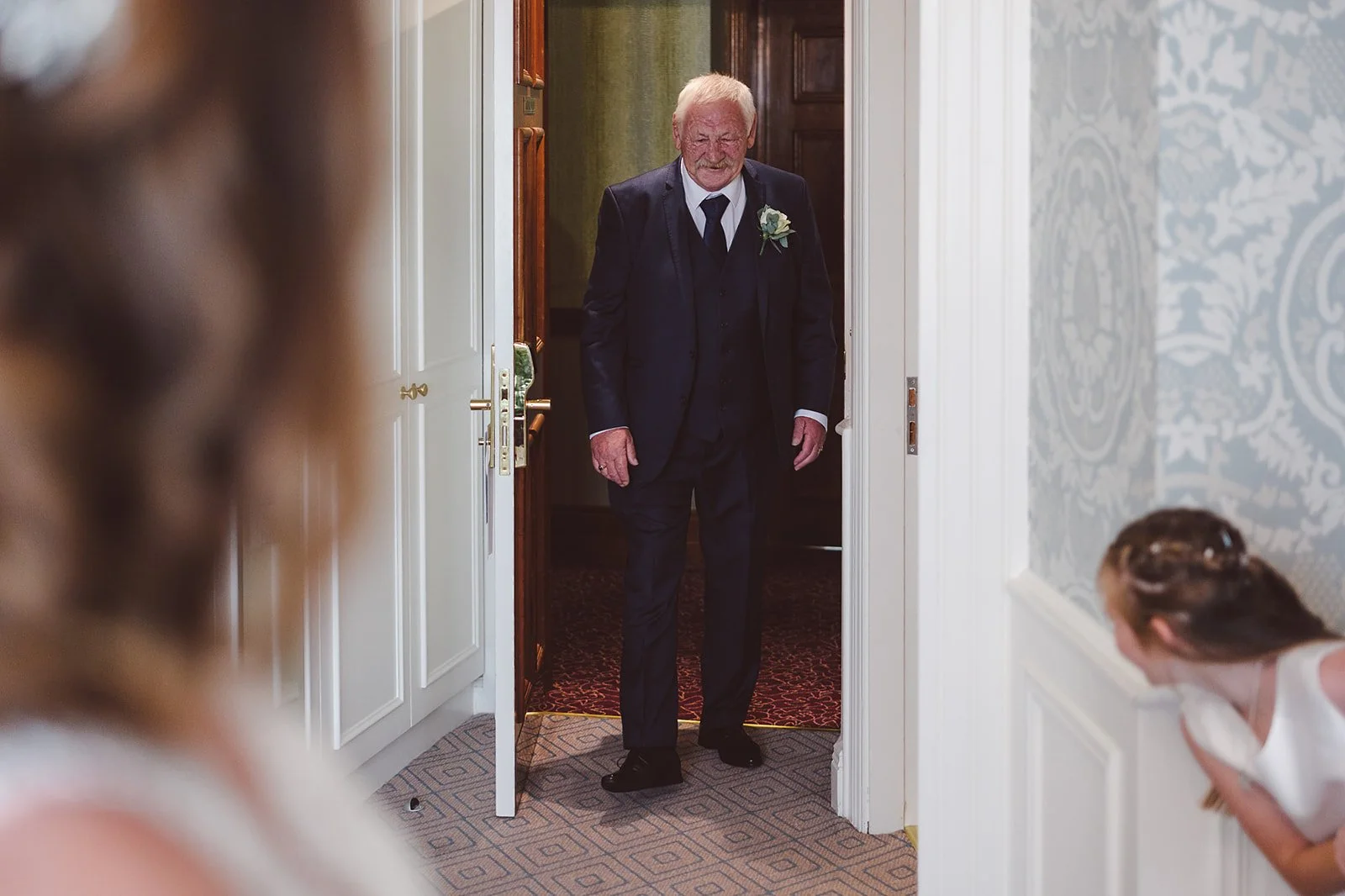 An elderly man in a dark suit with a flower boutonniere stands in a doorway, smiling at two women who are looking at him.