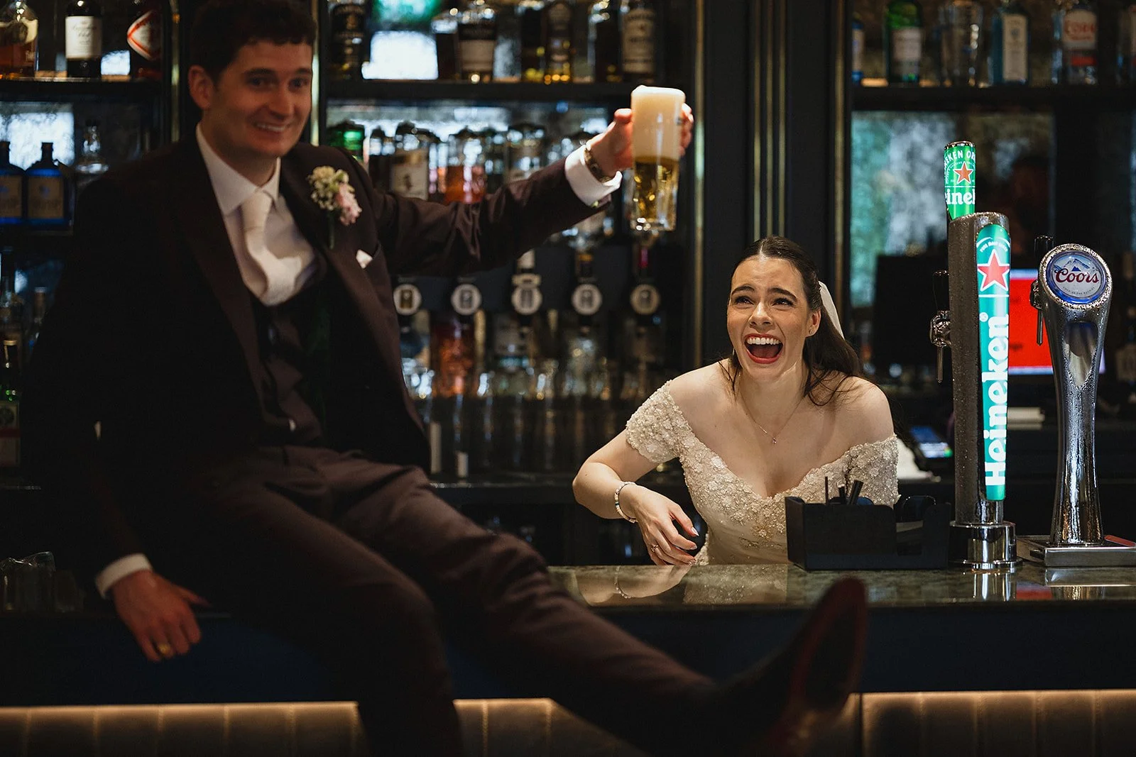 A man and woman at a bar, dressed in wedding attire. The man is holding a beer and smiling, while the woman is sitting on the bar counter, laughing.