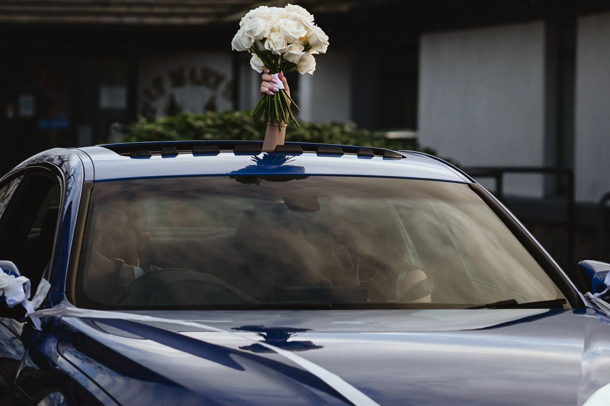 A person holding a bouquet of white roses out of the sunroof of a shiny black car during daylight.