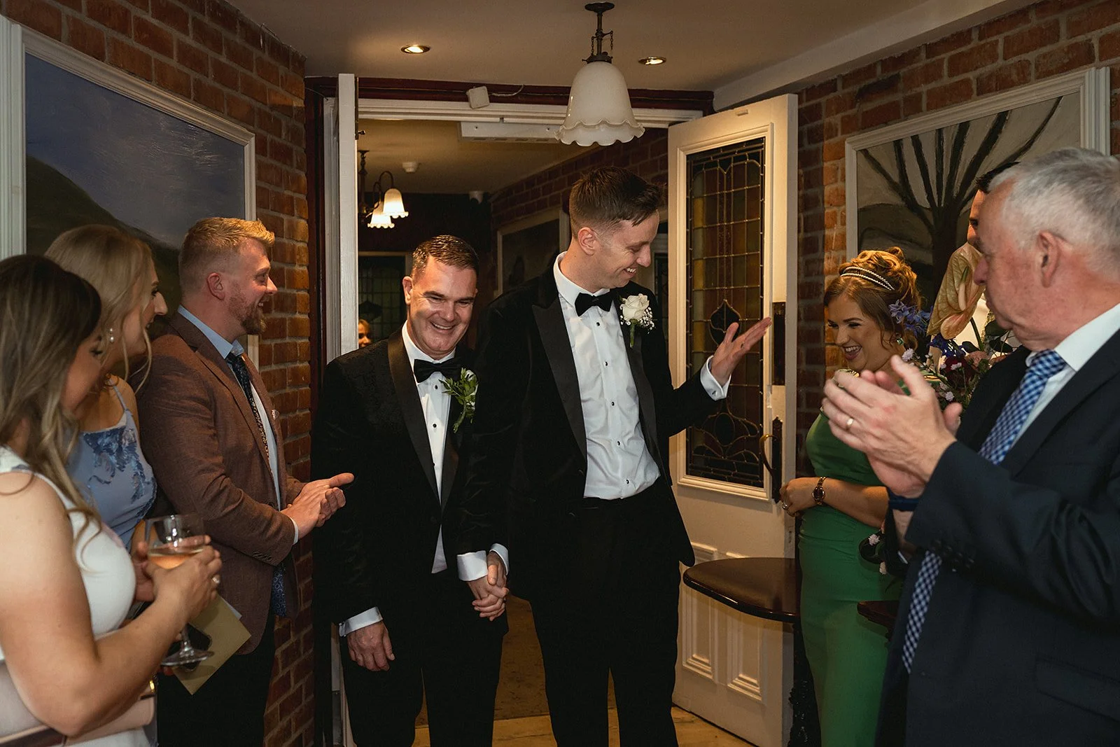 Two men in tuxedos holding hands and smiling at each other while surrounded by warmly dressed guests clapping and smiling inside a brick-walled house.
