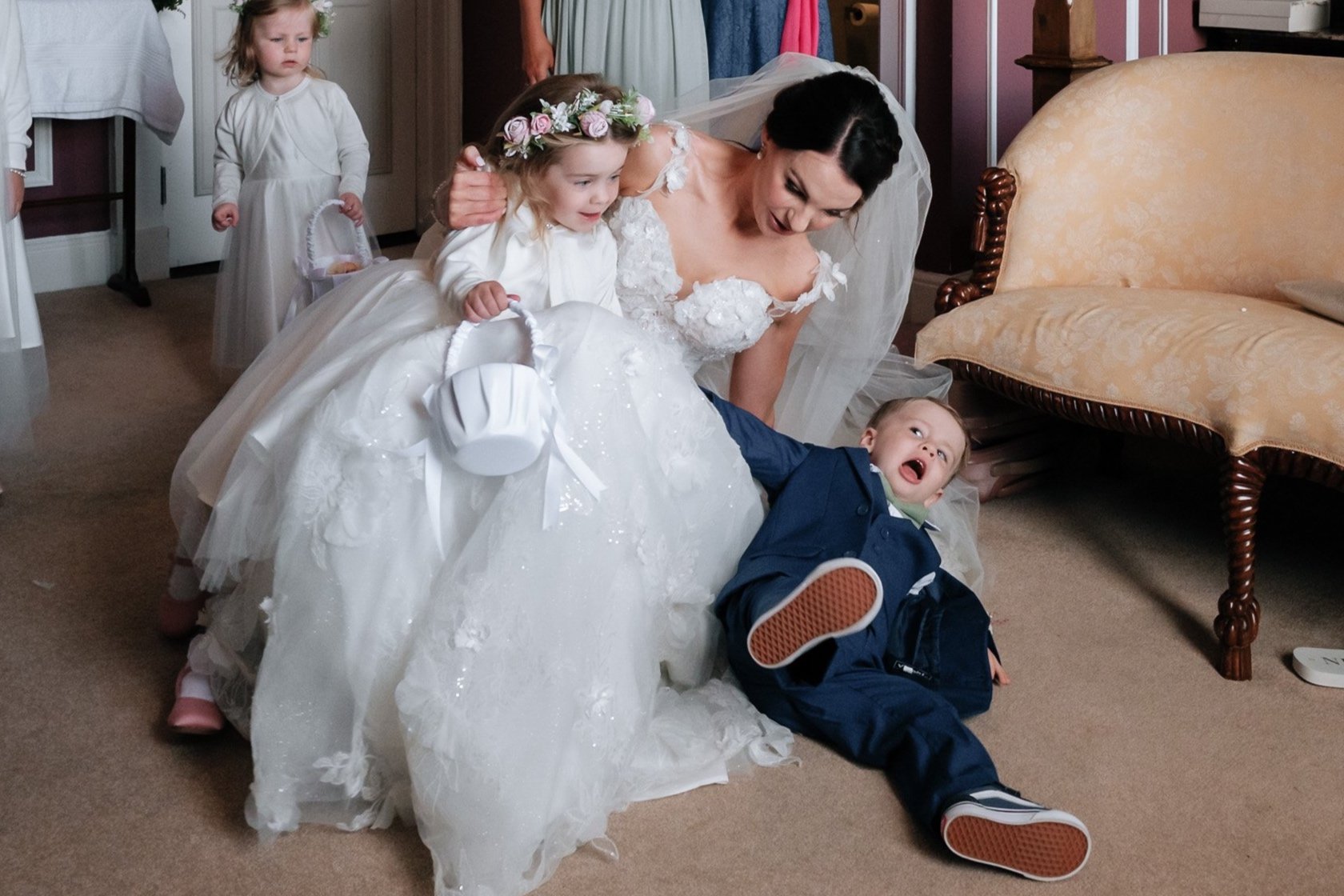 Bride with flower children in a dressing room. The bride is wearing a white gown and veil, holding a young girl in a white dress with a floral headband and a small white basket. A boy in a blue suit lies nearby. Another young girl in a similar white 