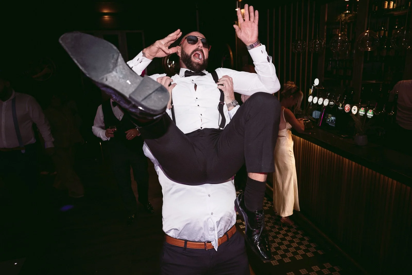 A man in a white shirt, black suspenders, and sunglasses is holding a woman in a white dress and black shoes in the air at a bar or nightclub. The man appears to be dancing or celebrating, with his mouth open and hands near his head. The woman is fac