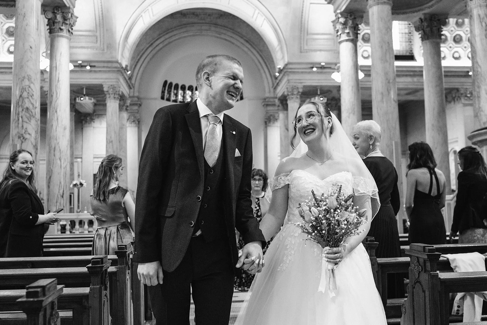 A couple gets married inside a grand church with tall marble columns, holding hands, smiling at each other. The bride wears a white wedding gown with lace details and a veil, and holds a bouquet of flowers. The groom is dressed in a suit with a tie. 