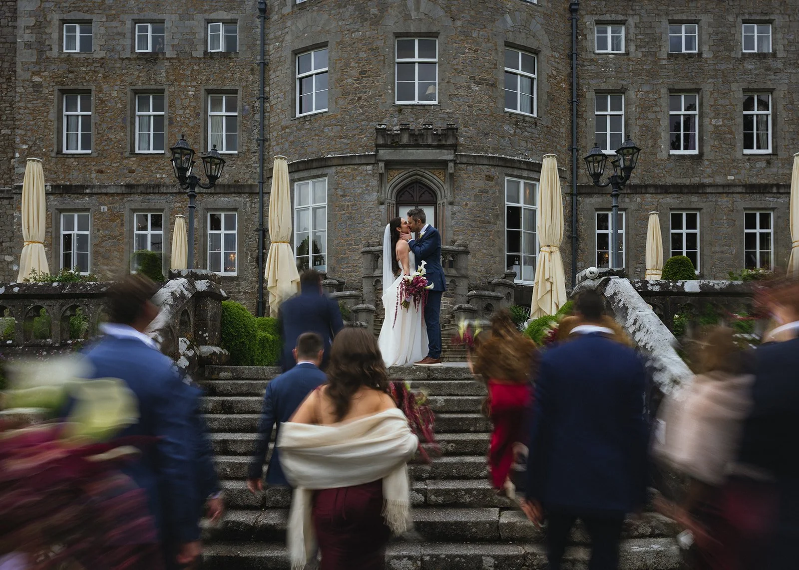 A wedding ceremony with a bride and groom kissing on the stairs outside a large stone building, surrounded by guests dressed in formal attire, at a venue with outdoor seating and umbrella-shaded tables.