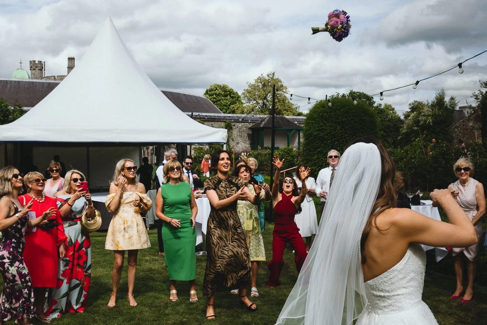 Wedding reception outdoors with guests watching the bouquet toss, bride in white dress with veil in foreground, bouquet flying in the air.