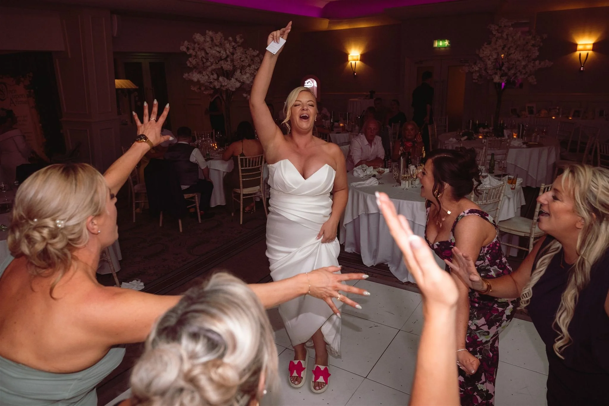 A woman in a white dress dancing and laughing with friends at a party or wedding reception.