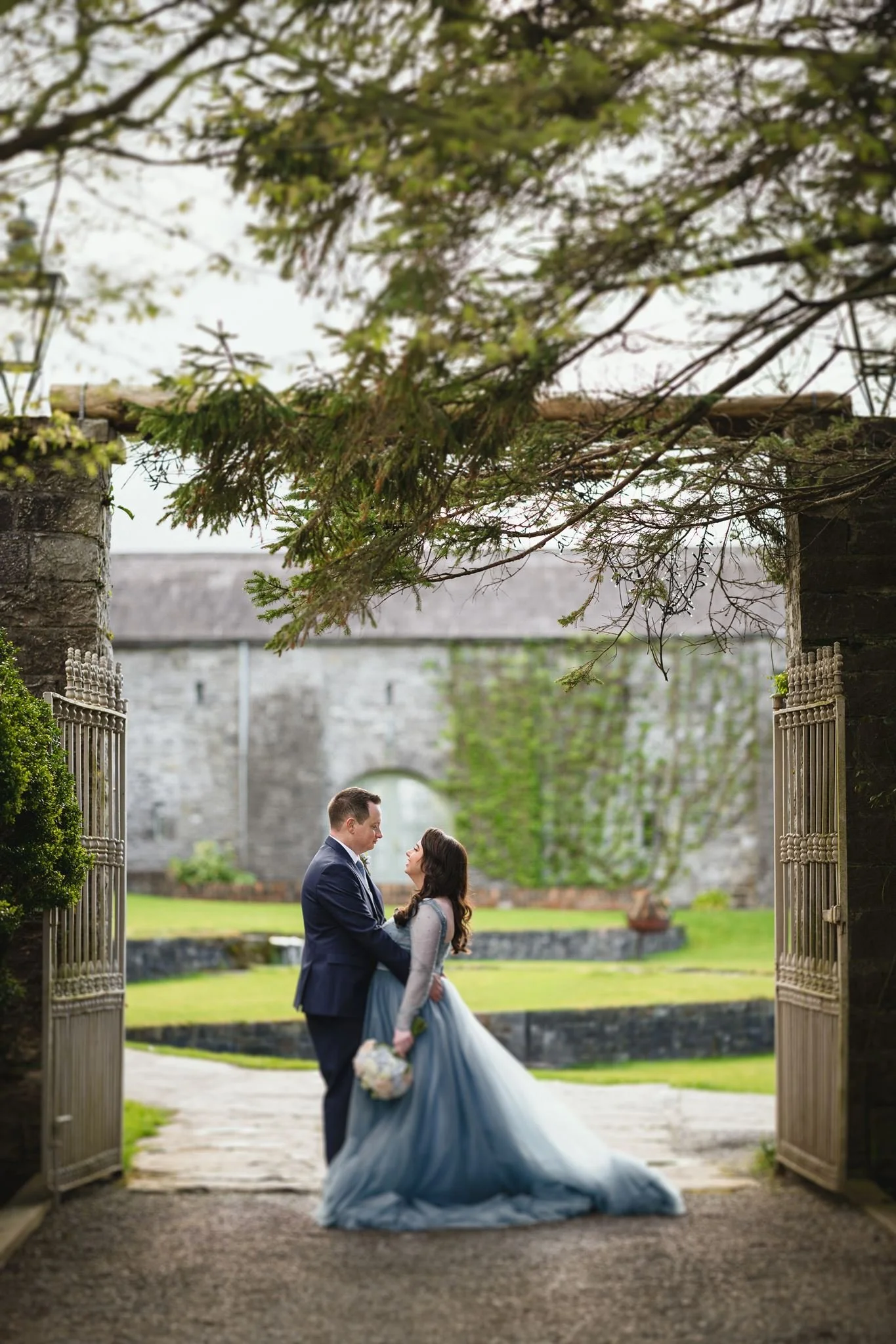 A bride and groom holding each other under an archway with wooden gates, greenery, and a historic stone building in the background.