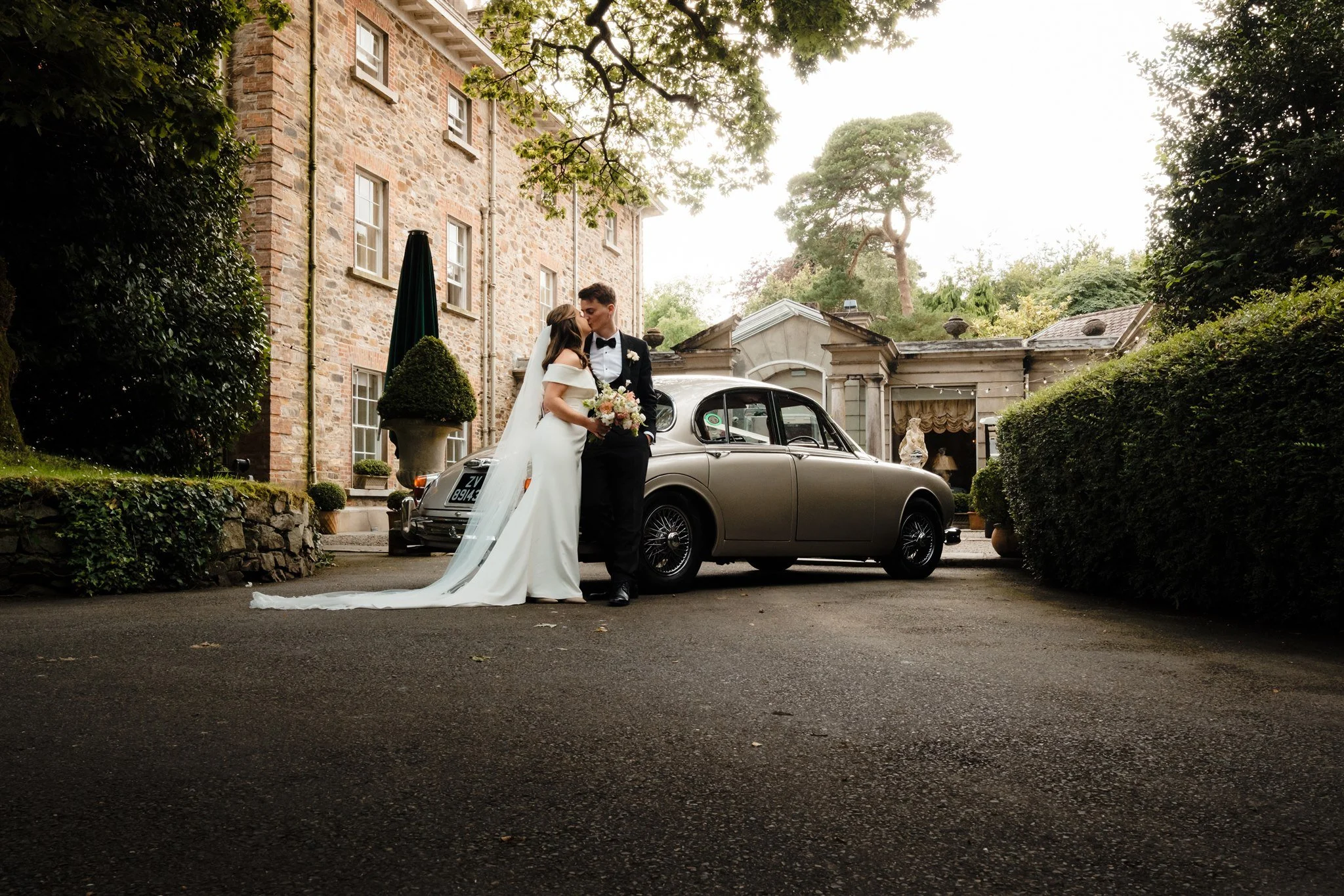 A newlywed couple sharing a kiss in front of a vintage car outside a stone building surrounded by greenery.