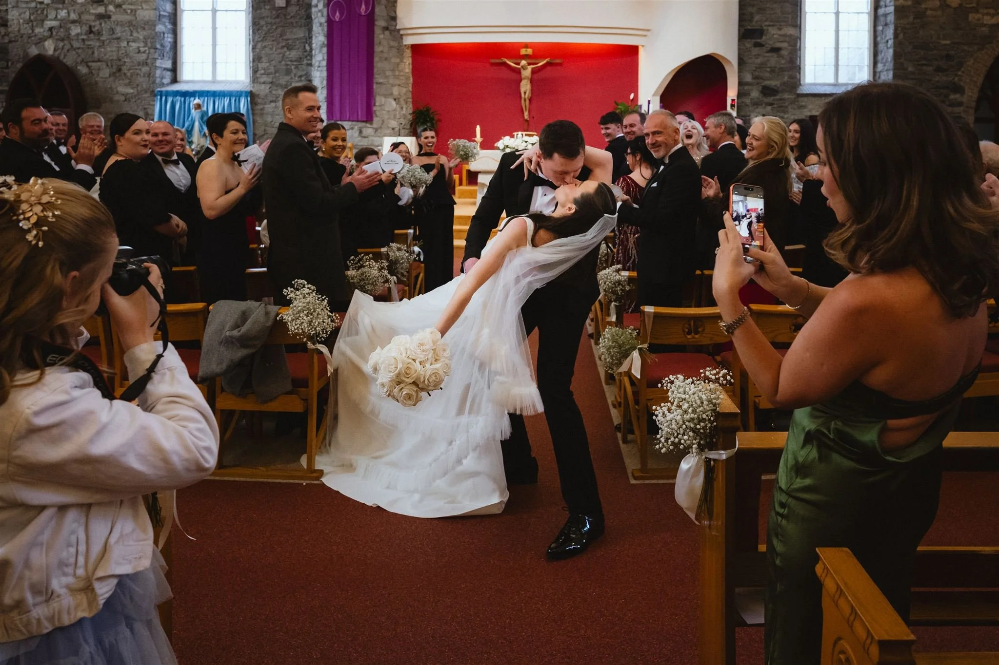 A bride and groom kissing during their wedding ceremony inside a church, with guests standing and taking photos. The church has stone walls and a red background with a crucifix above the altar.