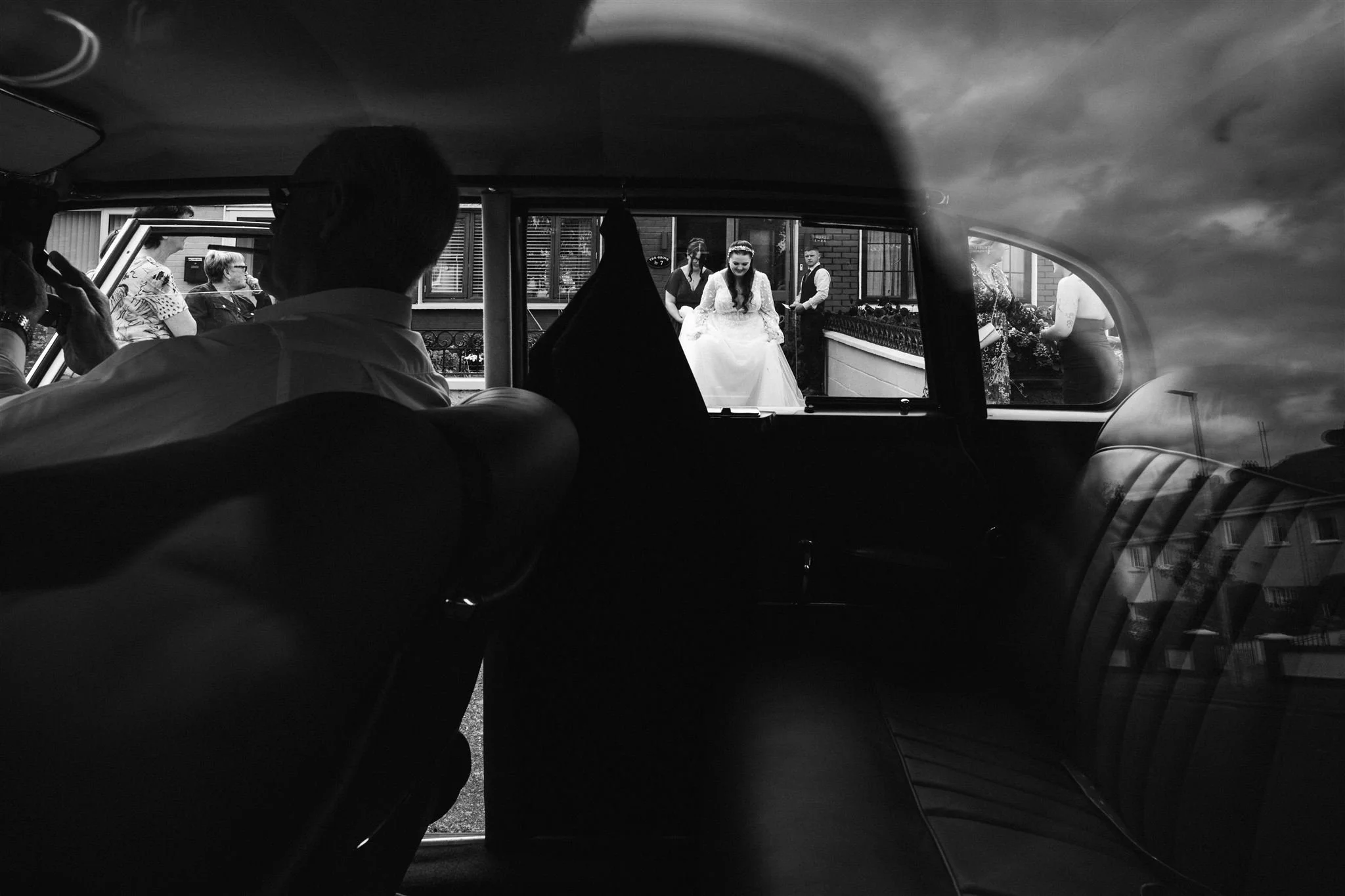 Black and white photo taken from inside a car showing the bride being dropped off outside a building, with people gathered outside.