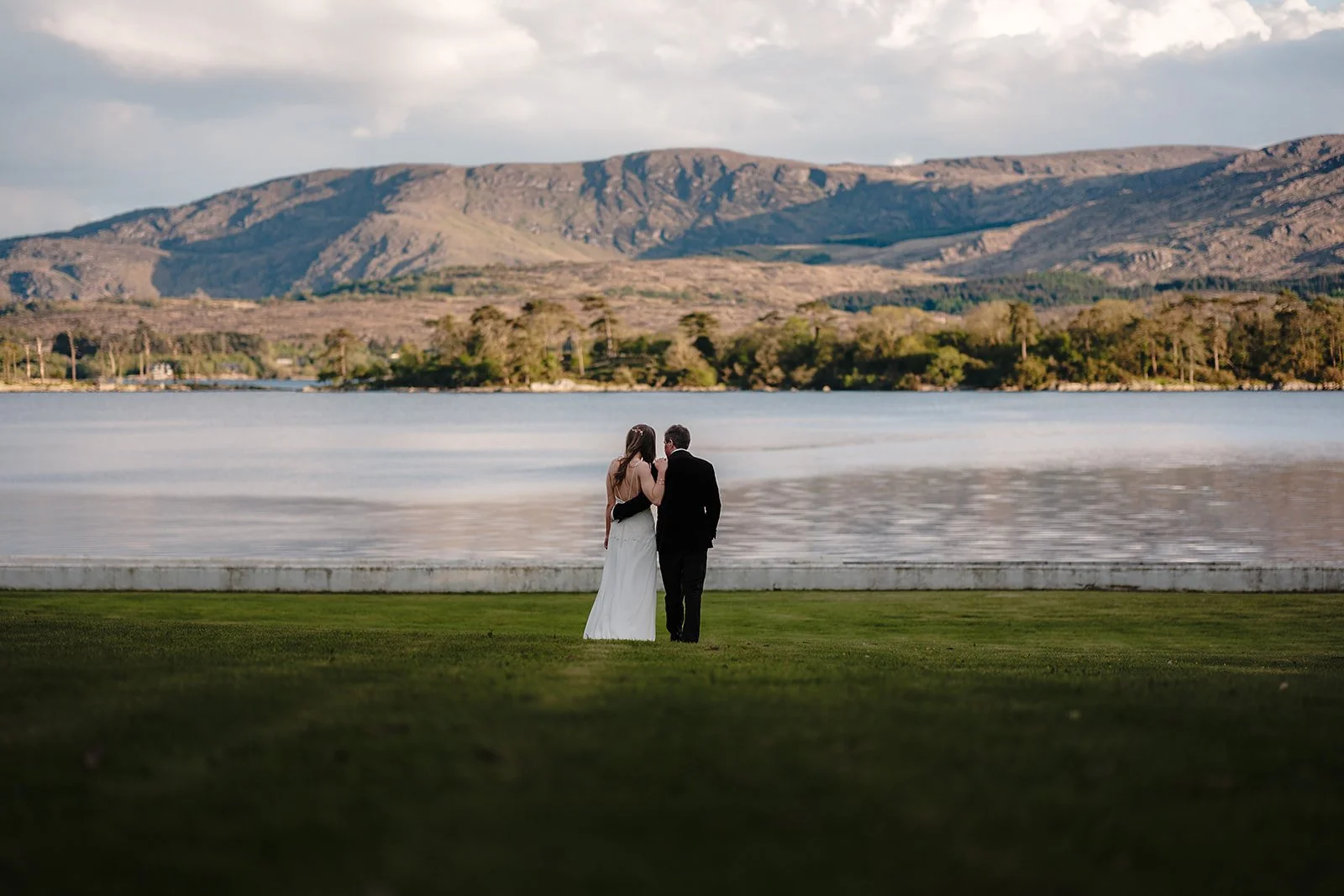 A bride and groom standing together by a lakeside, with mountains in the background, during sunset.