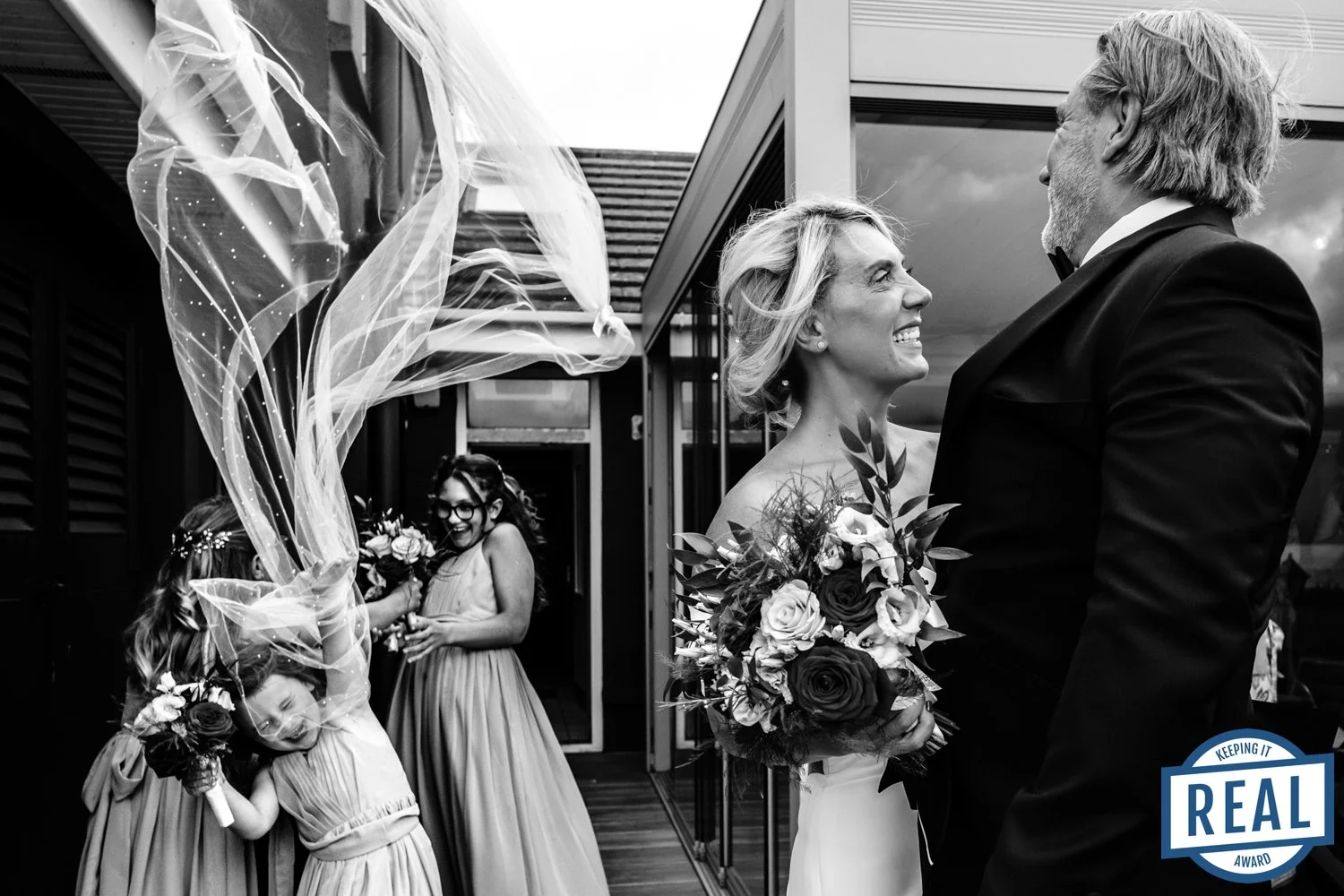 Black and white photo of a wedding couple smiling at each other, holding bouquets, with a woman and two children in matching dresses celebrating with flowers in the background.