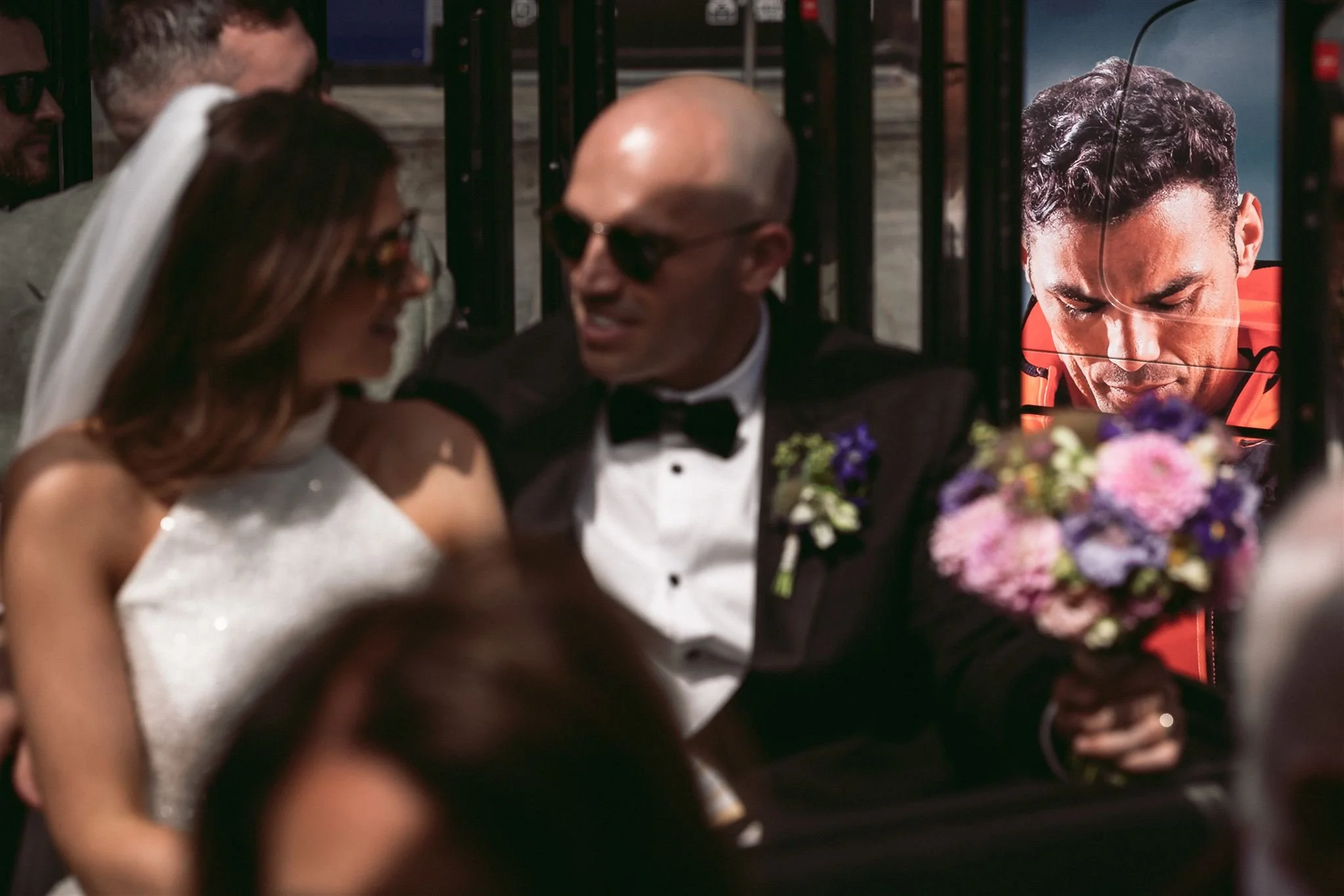 A wedding scene with a bride in a white dress and a groom in a tuxedo sharing a kiss, with a man holding a bouquet of flowers and a large photo of the groom in the background.