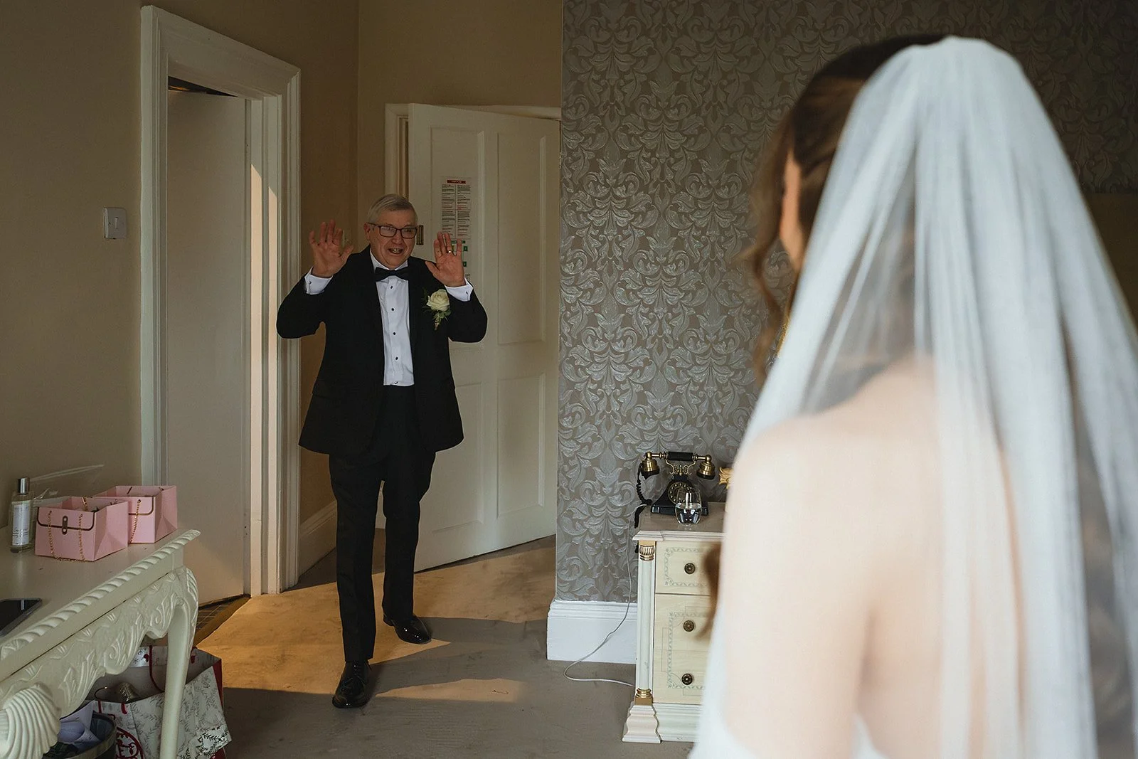 A groom in a tuxedo with a boutonniere smiles and raises his hands as he sees the bride, whose back is to the camera, wearing a white veil.