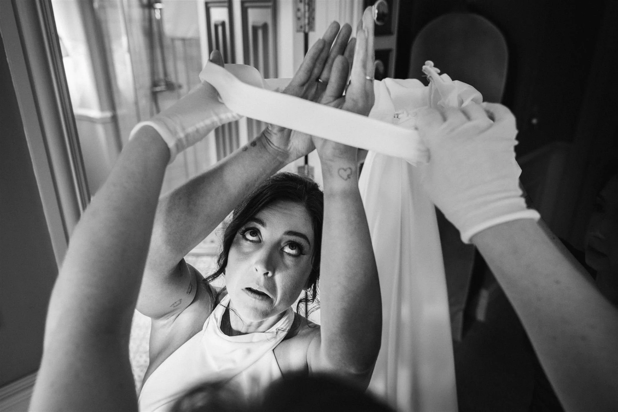 A woman looks up at a wedding dress being hung or adjusted by others, with her hands above her head. Black and white photo, behind a window and some furniture.