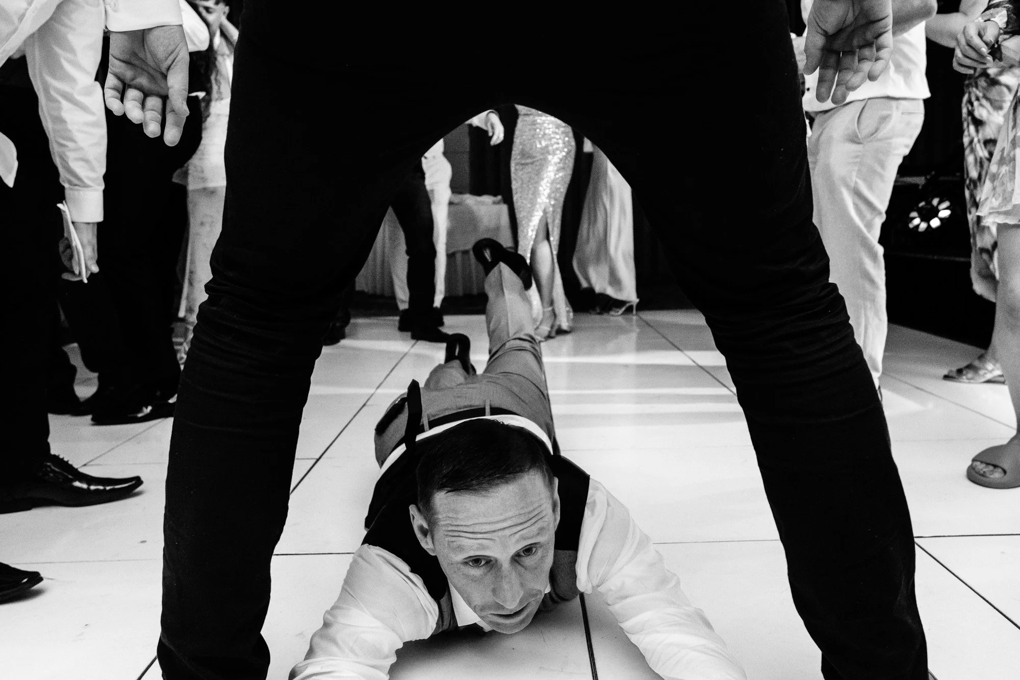 Black and white image of a person in formal attire crawling on the floor at a party, surrounded by standing people.
