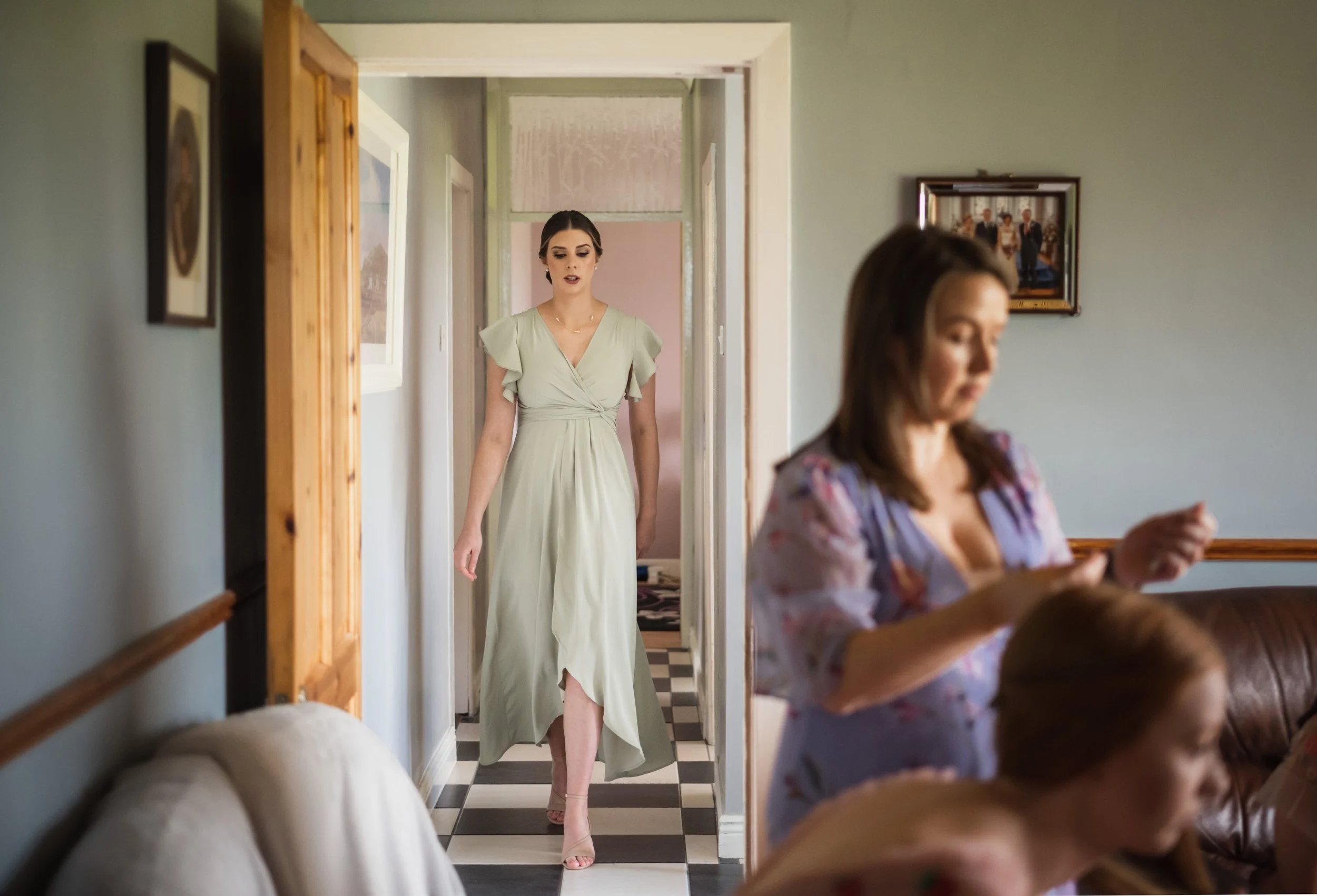 A woman in a light green dress walking through a doorway, with another woman in the foreground using a phone or camera, in a room with framed pictures on the walls and a black-and-white checkered floor.