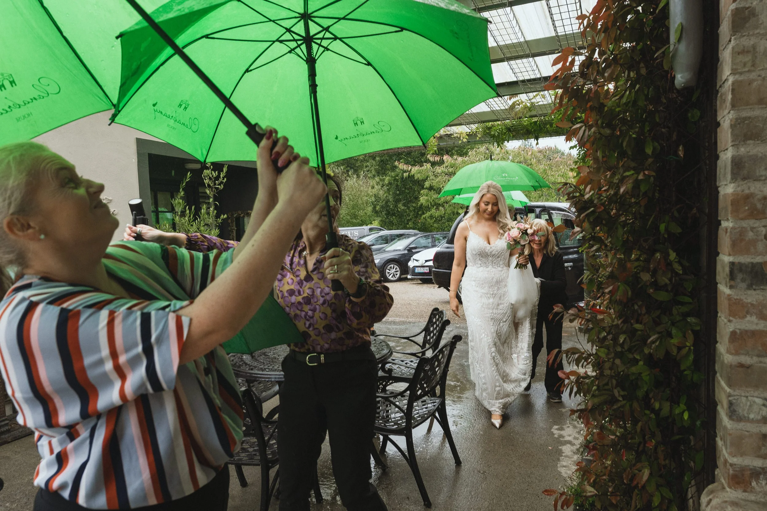 A bride in a white wedding dress holding a bouquet, walking outdoors on a rainy day under a green umbrella held by a guest. Other guests also hold umbrellas, and cars are visible in the background.