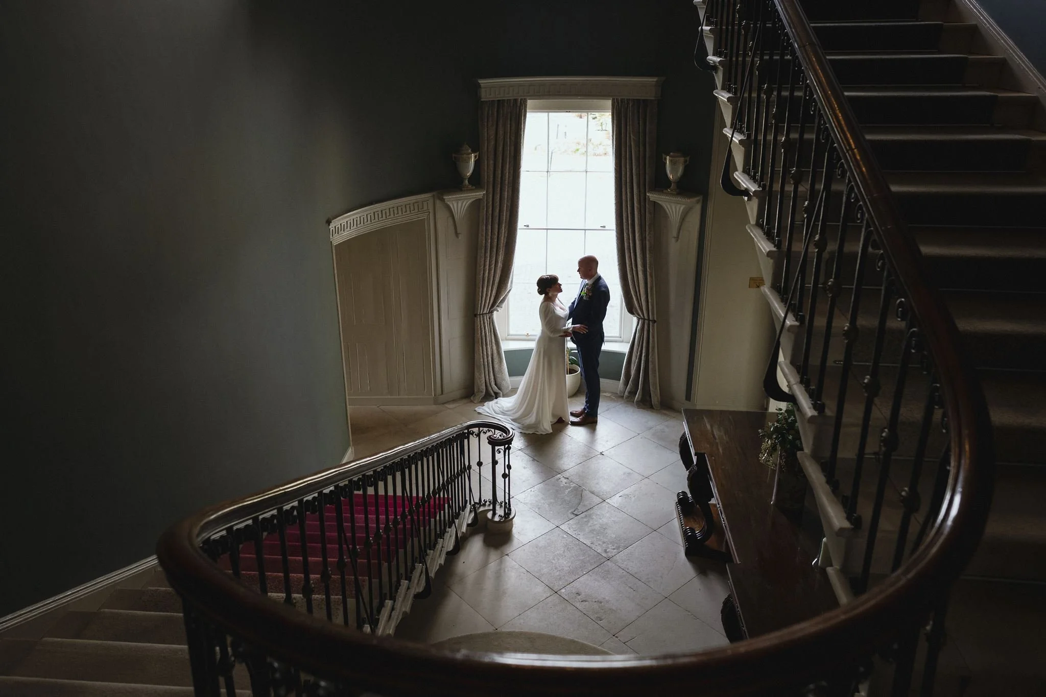A bride and groom standing together in front of a large window with curtains, inside a grand staircase area with elegant decor.