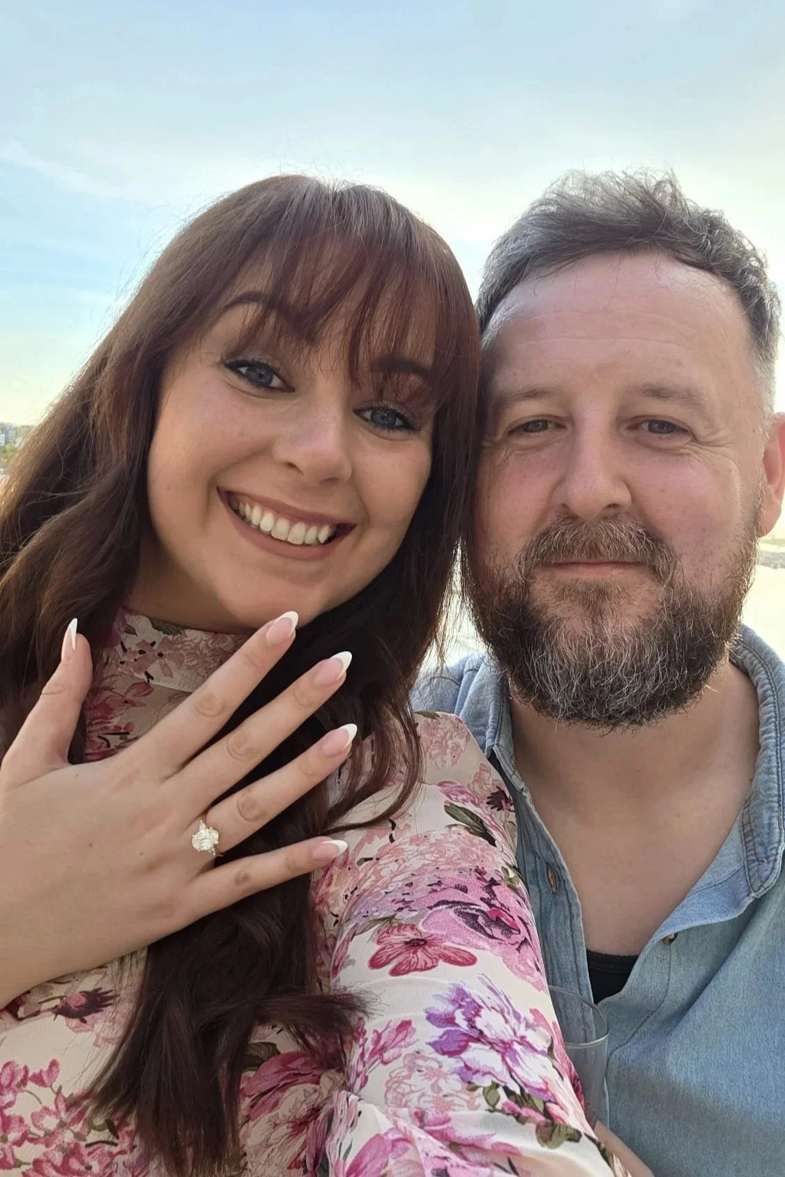 A smiling woman showing off her engagement ring while standing next to a man, both taking a selfie outdoors with a blue sky in the background.