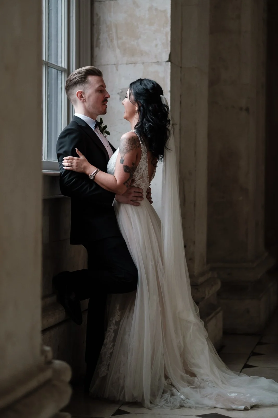 Bride and groom in wedding attire in a historic building with stone walls and large windows, standing close and looking into each other's eyes.