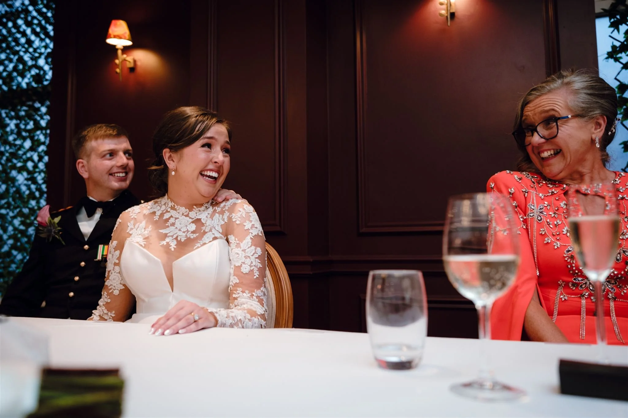 A bride and groom sitting at a wedding reception table, laughing and talking with an older woman who is also smiling, in a warmly lit room with dark wood walls.
