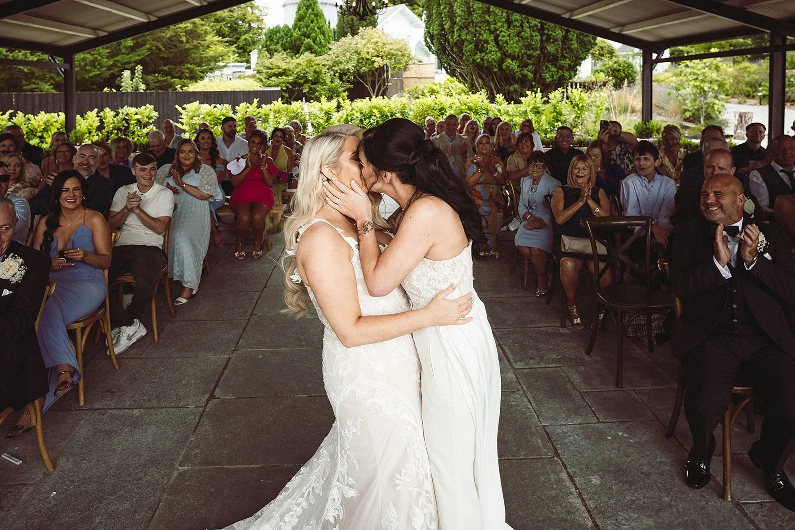 Two women, wearing white wedding dresses, are kissing during a wedding ceremony with guests seated around them, some clapping and smiling.