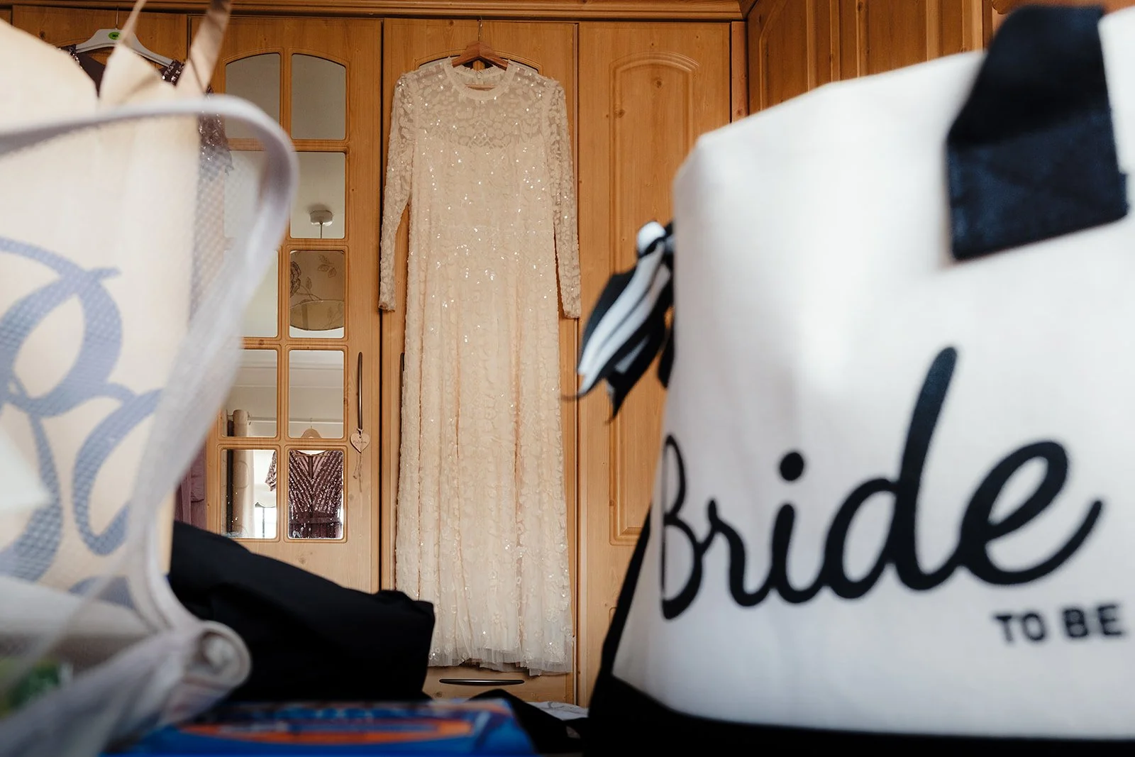 A white wedding dress with sequins hanging on a wooden wardrobe, framed by two shopping bags in the foreground, one labeled 'bride to be'.