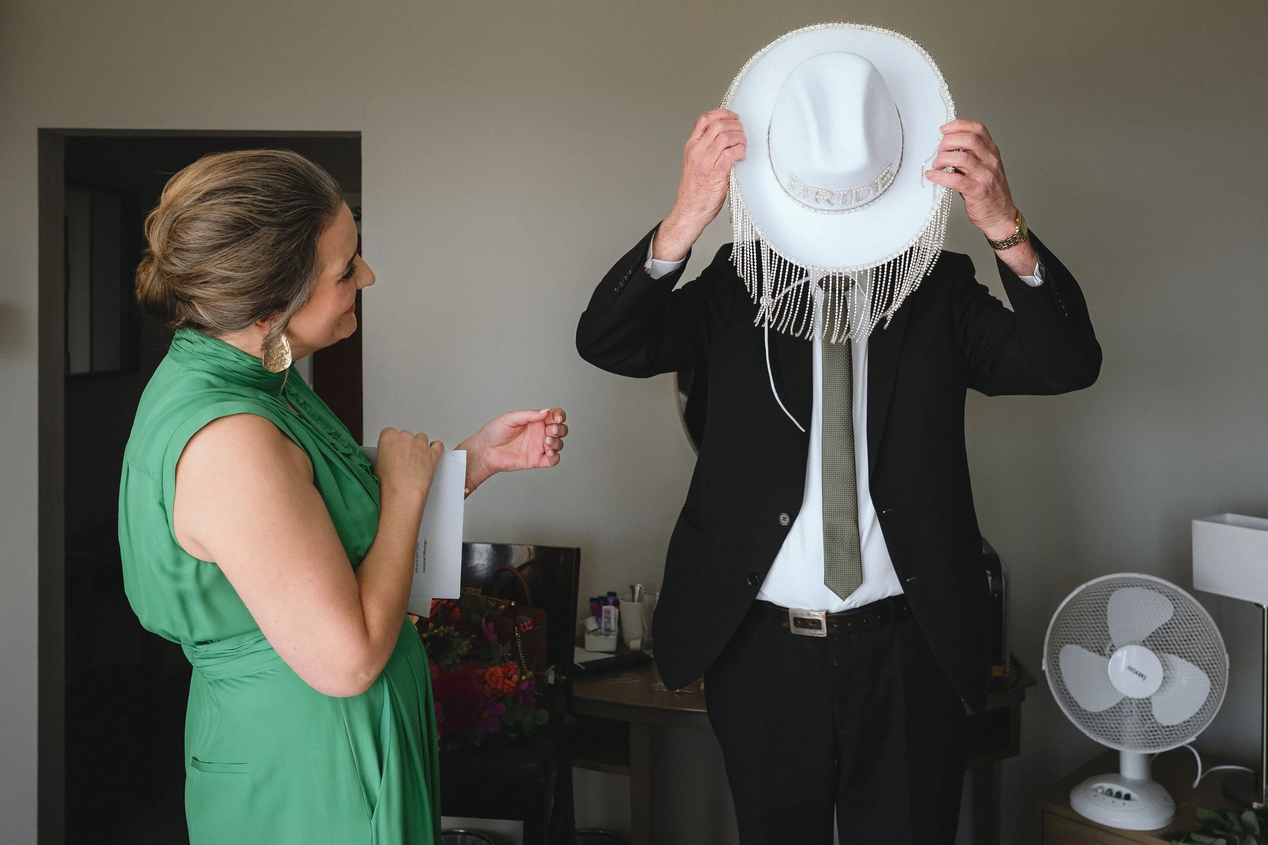 A woman in a green dress watching a man in a black suit trying on a white cowboy hat with a fringe and the word 'BRIDE' on it, inside a room with a fan on a table.