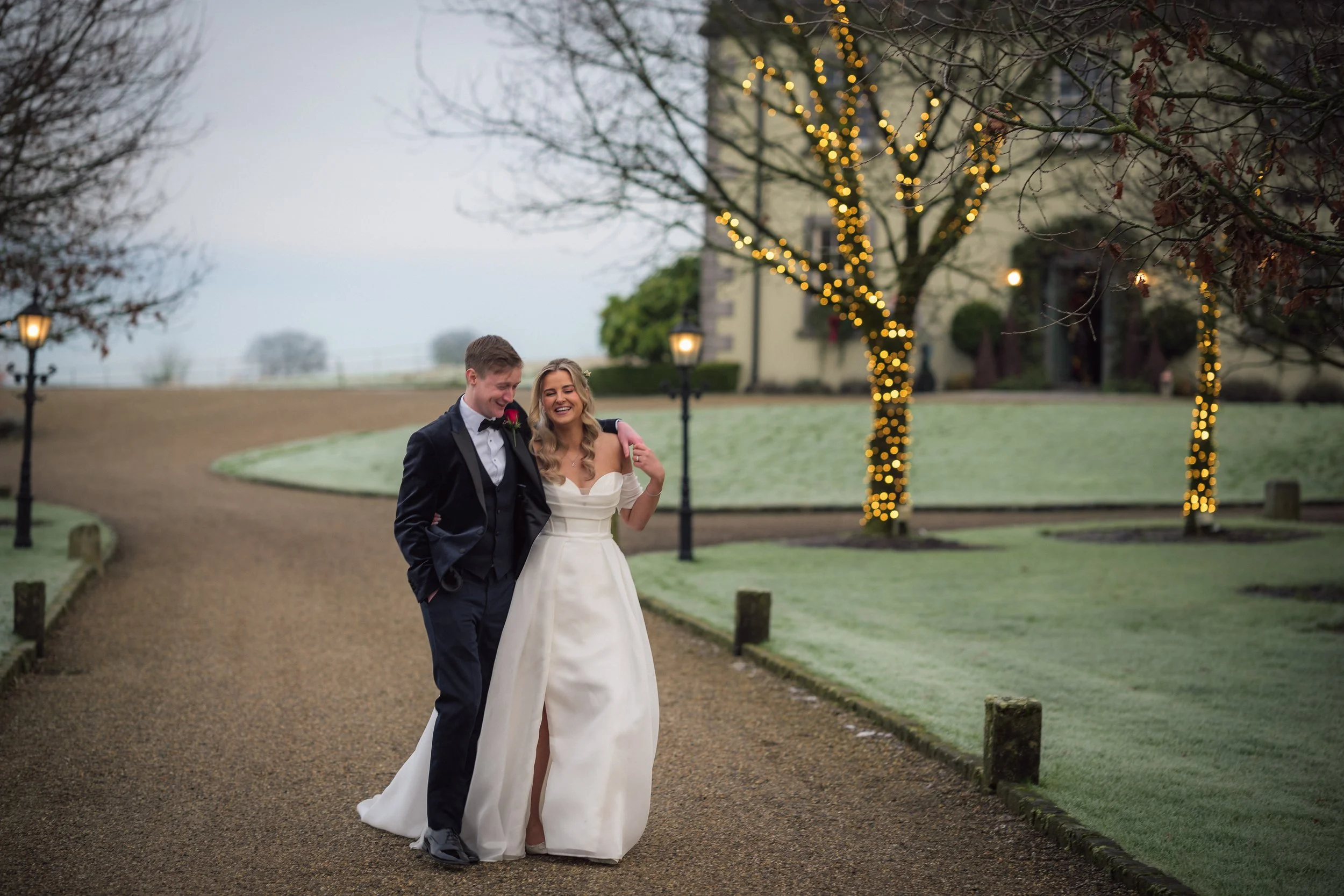 Smiling couple in formal attire walking on a path with trees wrapped in string lights.