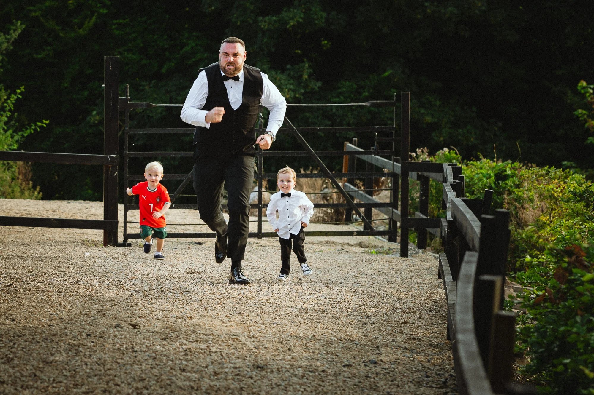 A man in formal attire running outdoors with two young boys, one in a tuxedo shirt and the other in a sports jersey, on a gravel pathway with a black wooden fence and greenery in the background.