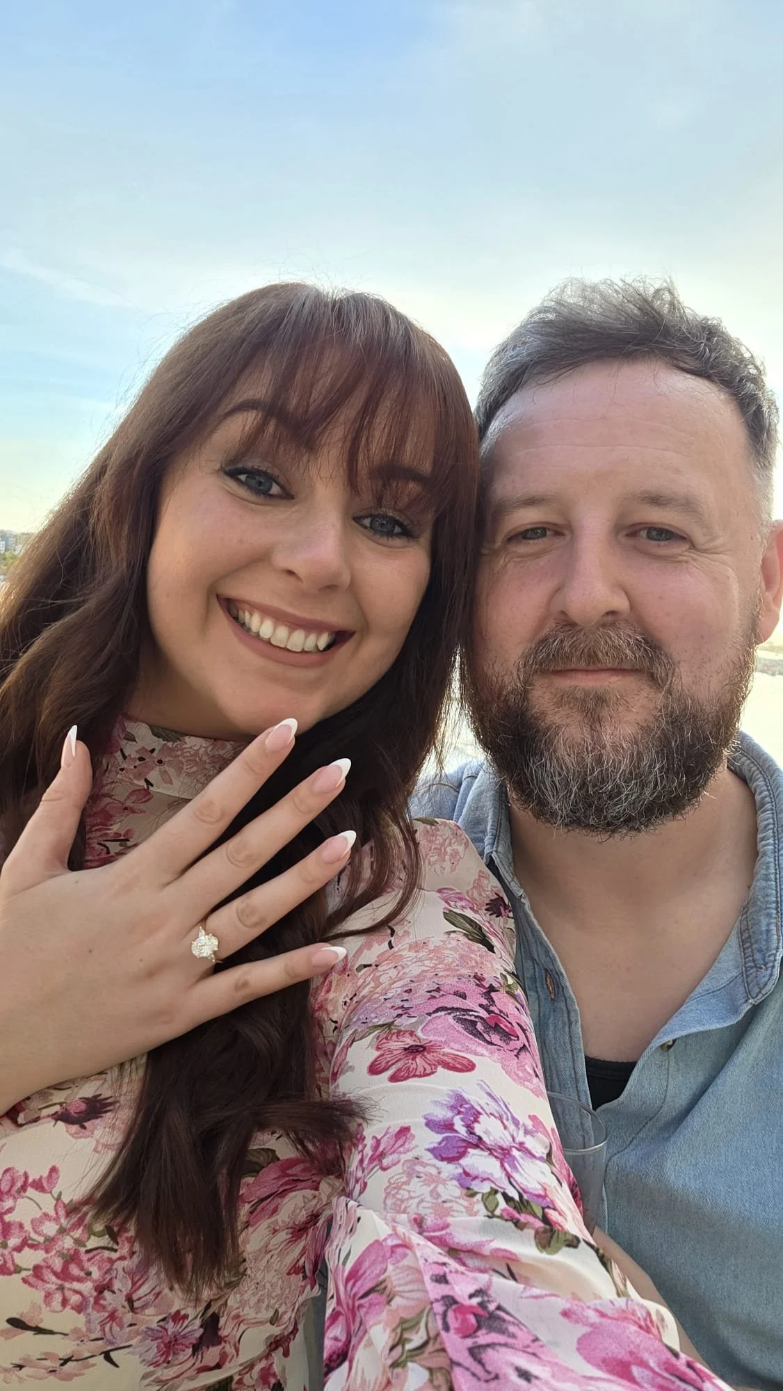 A smiling woman showing off a diamond ring on her finger next to a bearded man, outdoors with a blue sky in the background.